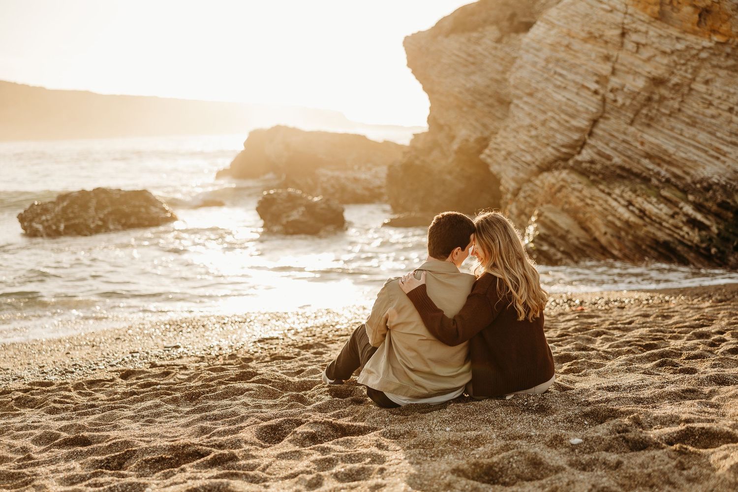 Couple sits together on beach at sunset with dramatic coastal rock formations.