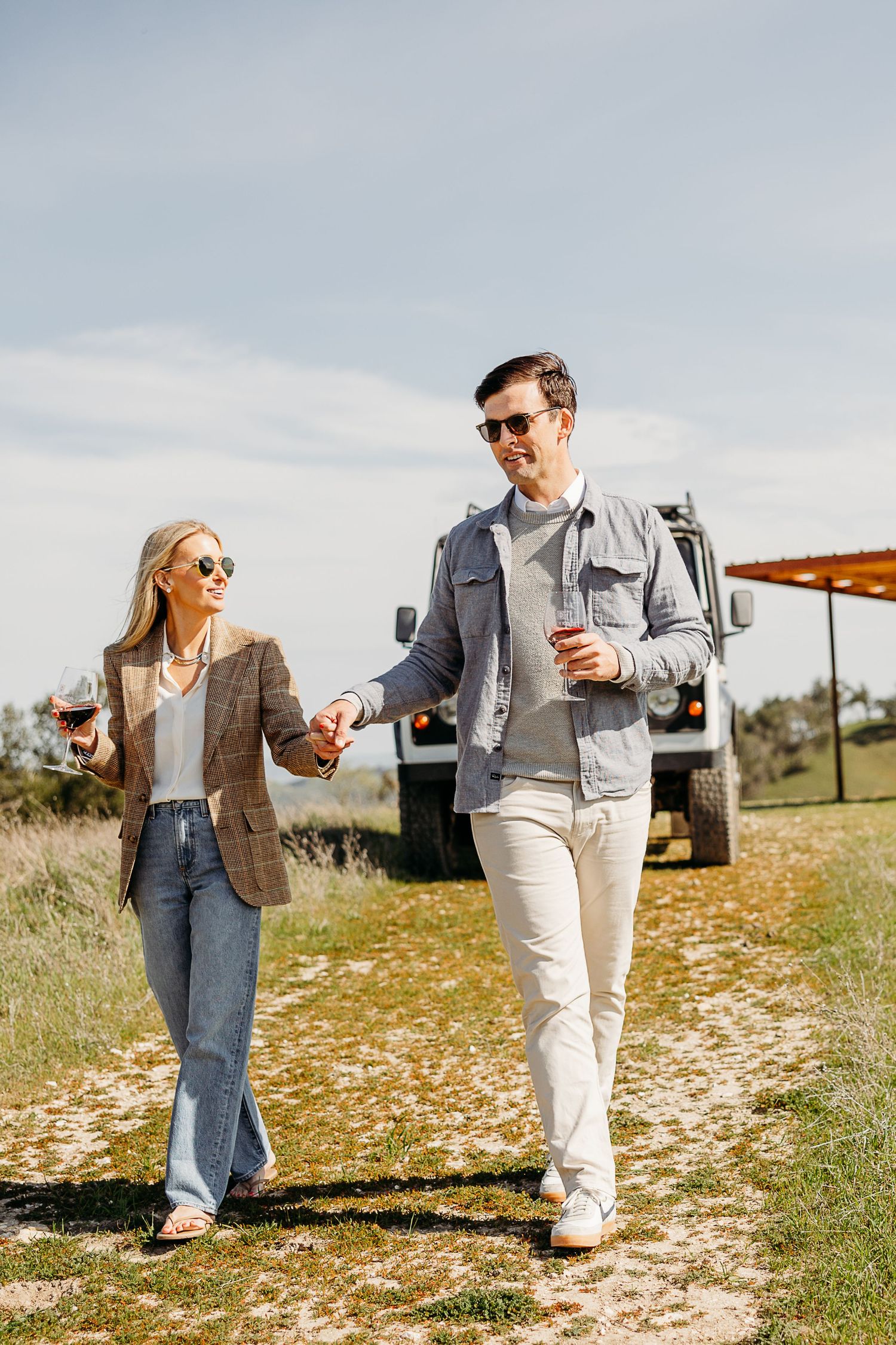 Couple walking along a hilltop path while holding wine glasses.