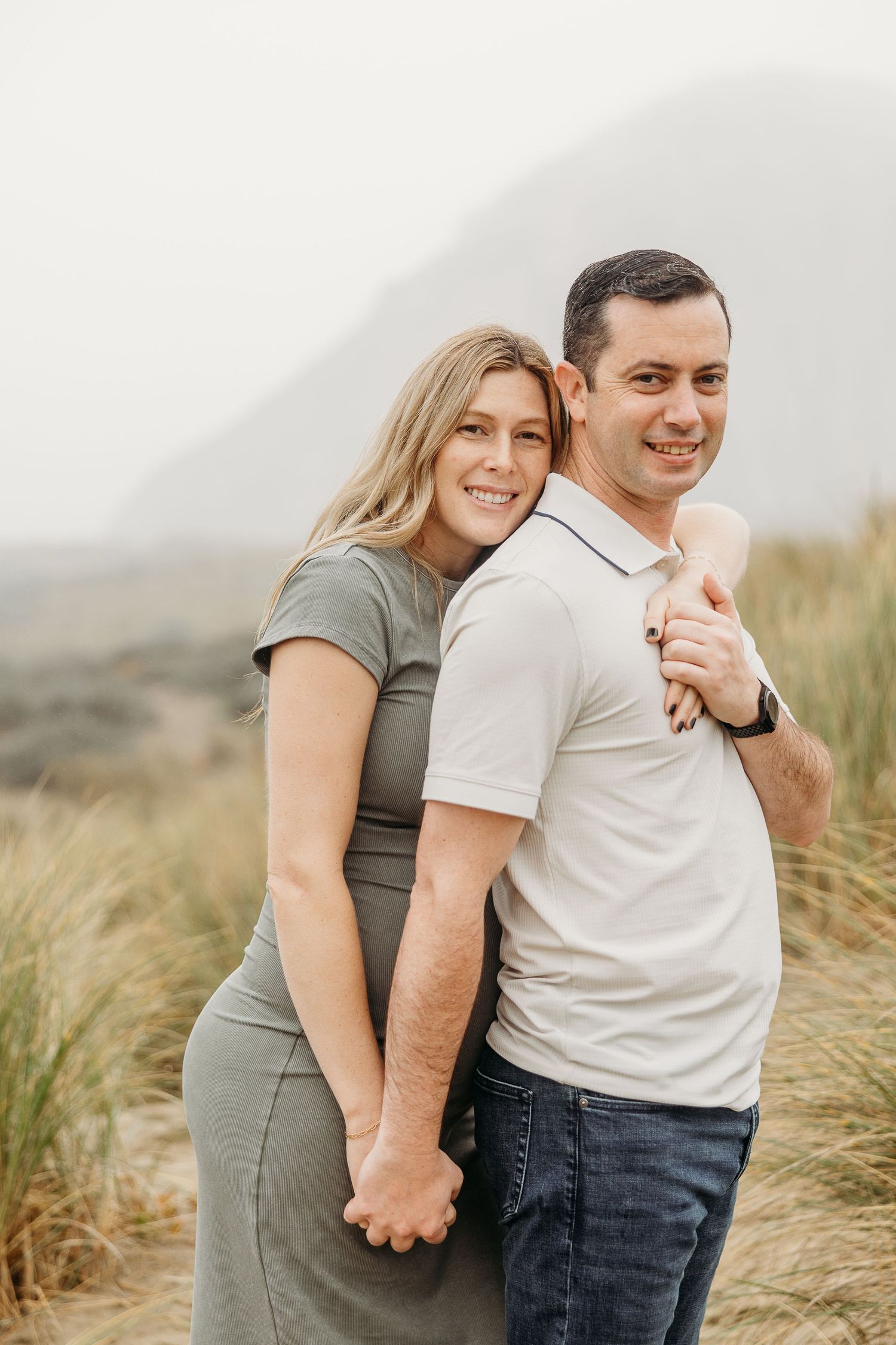 Joyful couple poses together against coastal landscape in maternity photoshoot.