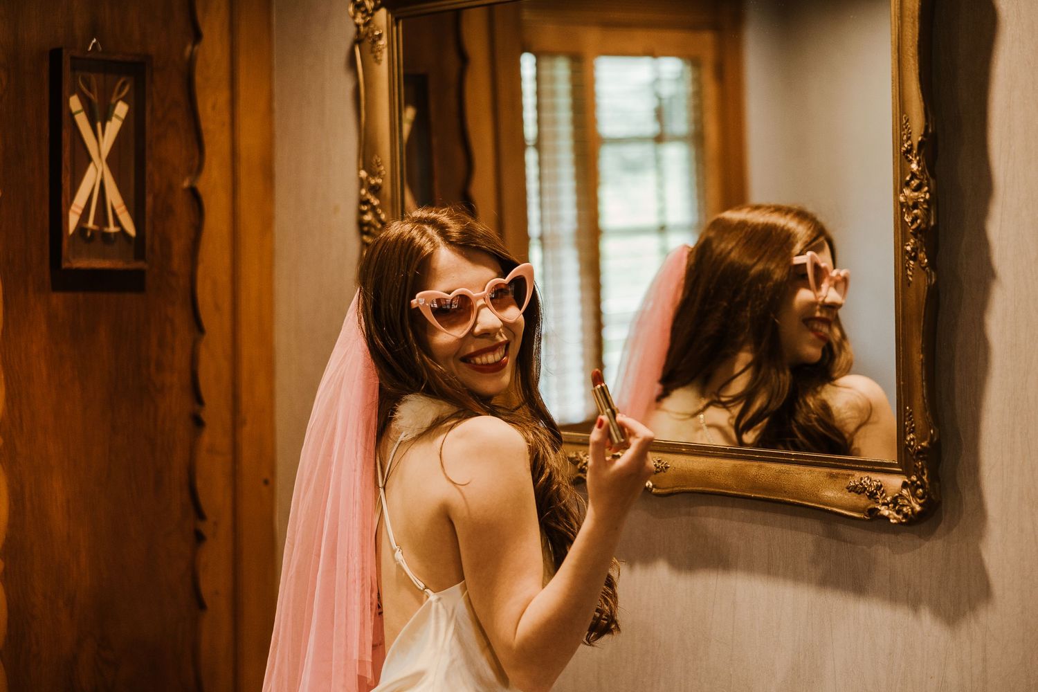 Person in a bridal veil smiles while getting ready in front of an ornate gold mirror in a rustic wood-paneled room.