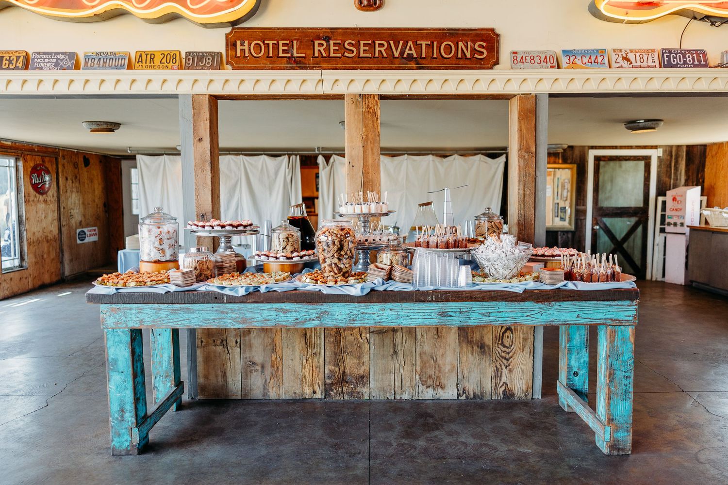 A rustic reception table with turquoise wood base displays vintage glassware and decor under a hotel reservations sign.