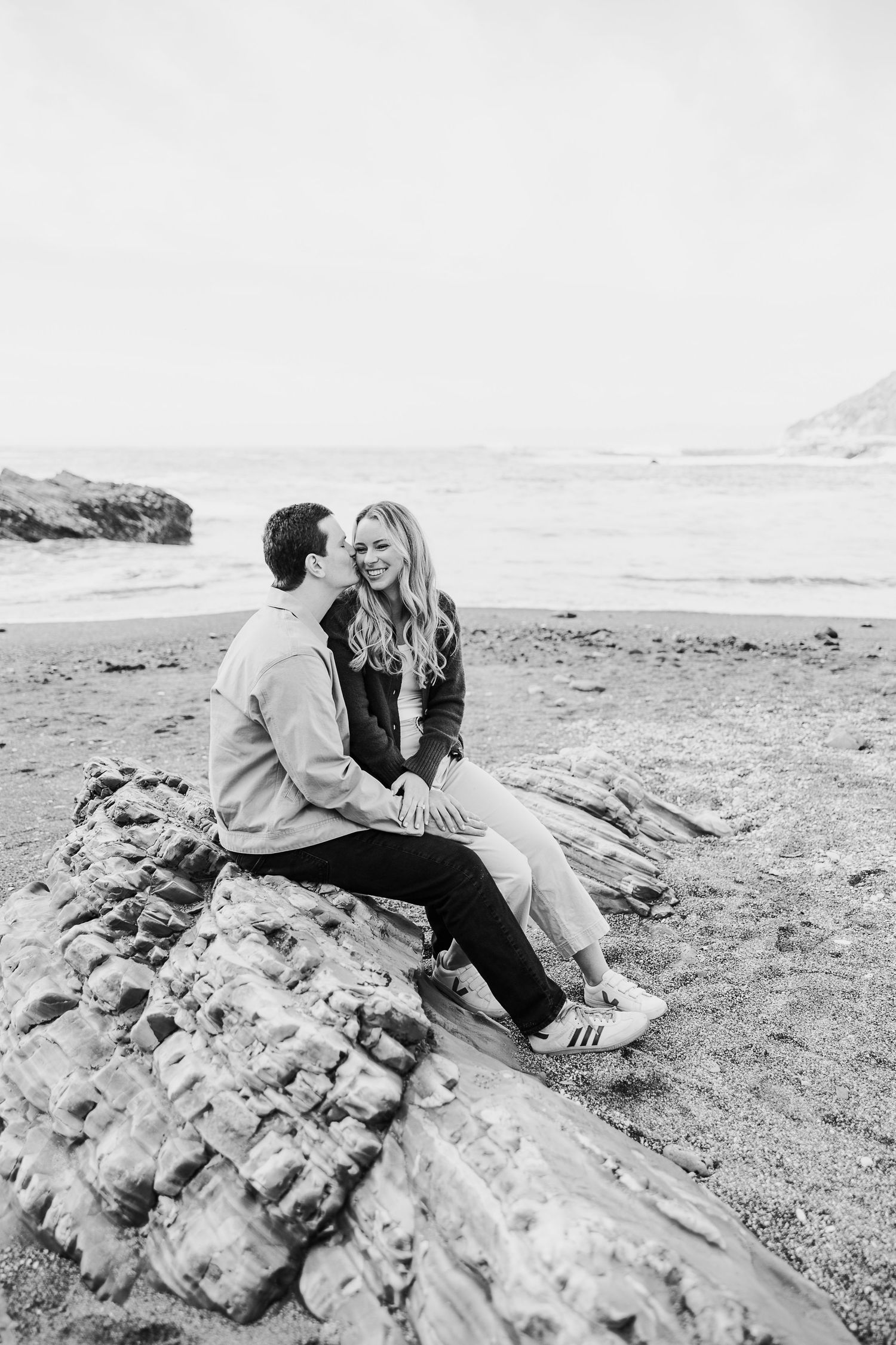 Two people share intimate moments while sitting on coastal rocks by the ocean.
