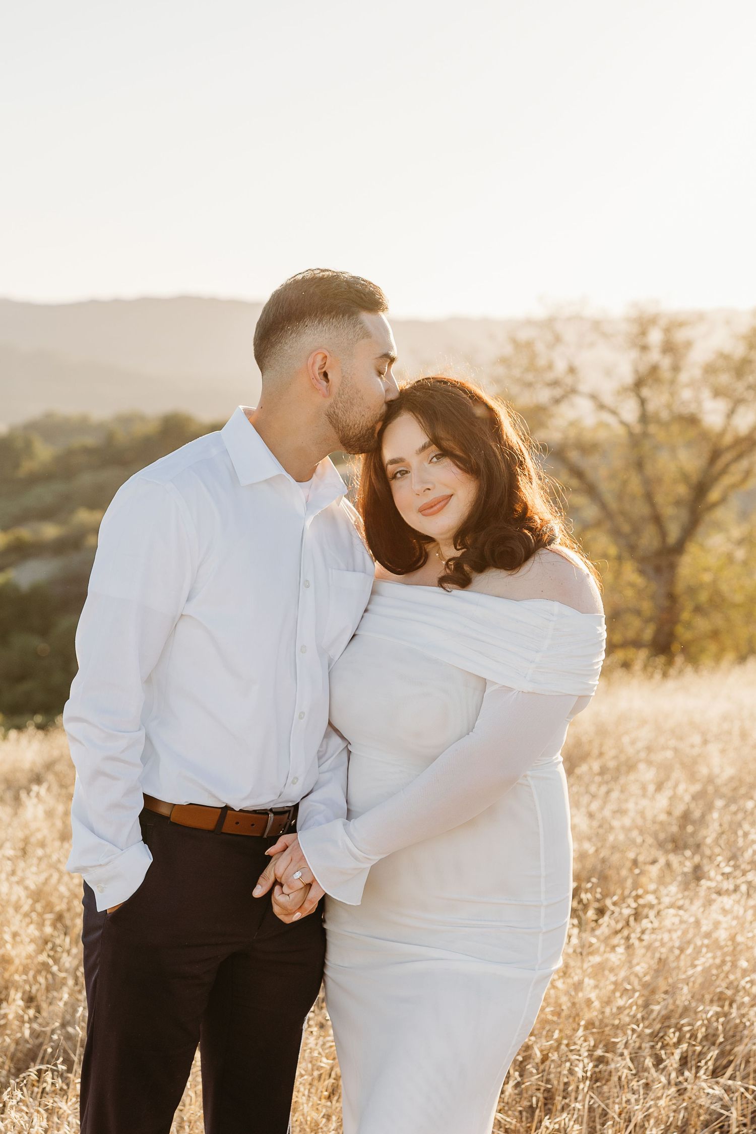 A couple in white and black outfits embrace lovingly in a sunlit field of golden grass during sunset.