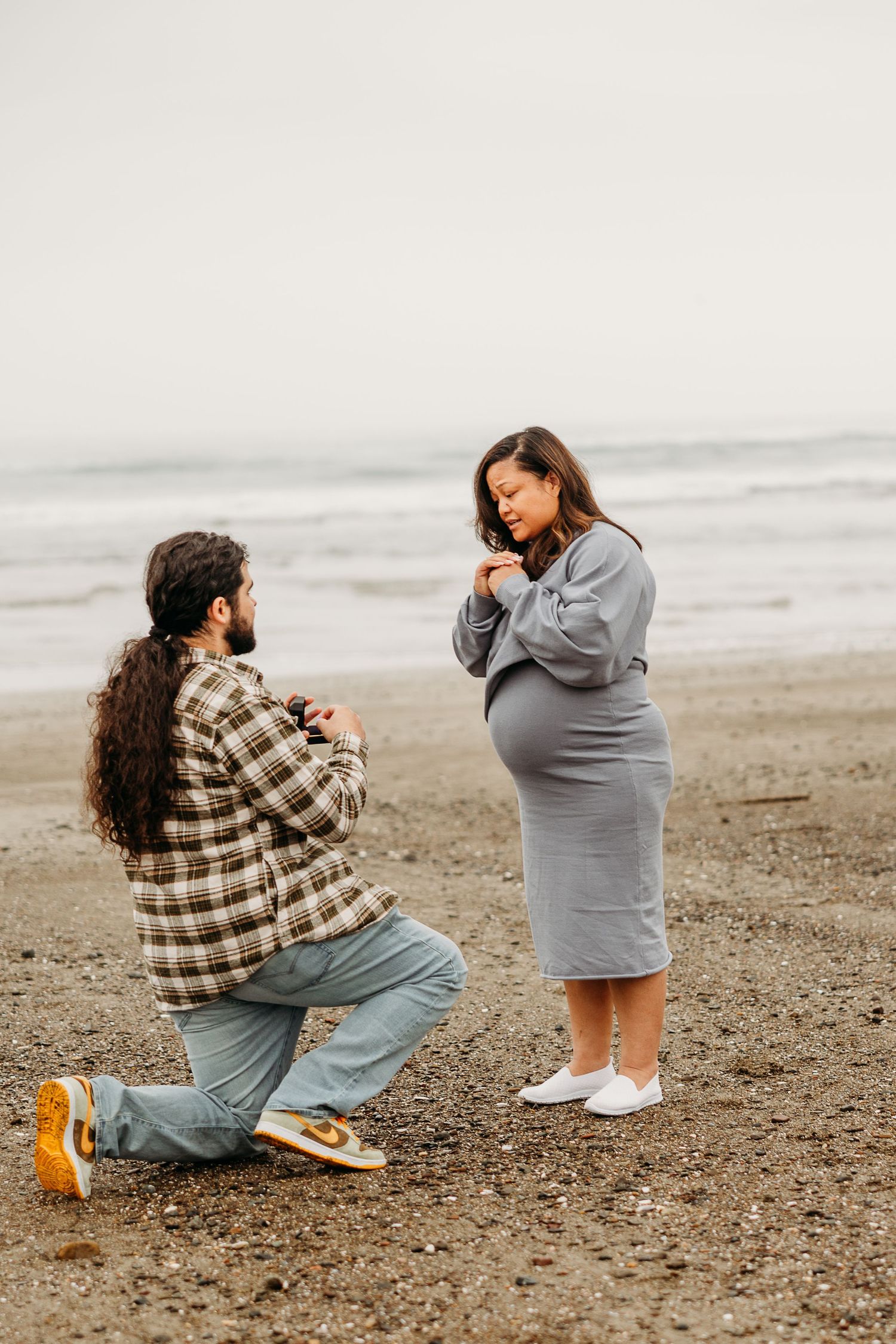 Couple sharing emotional moment during beach proposal with moody overcast sky and sand in muted tones.