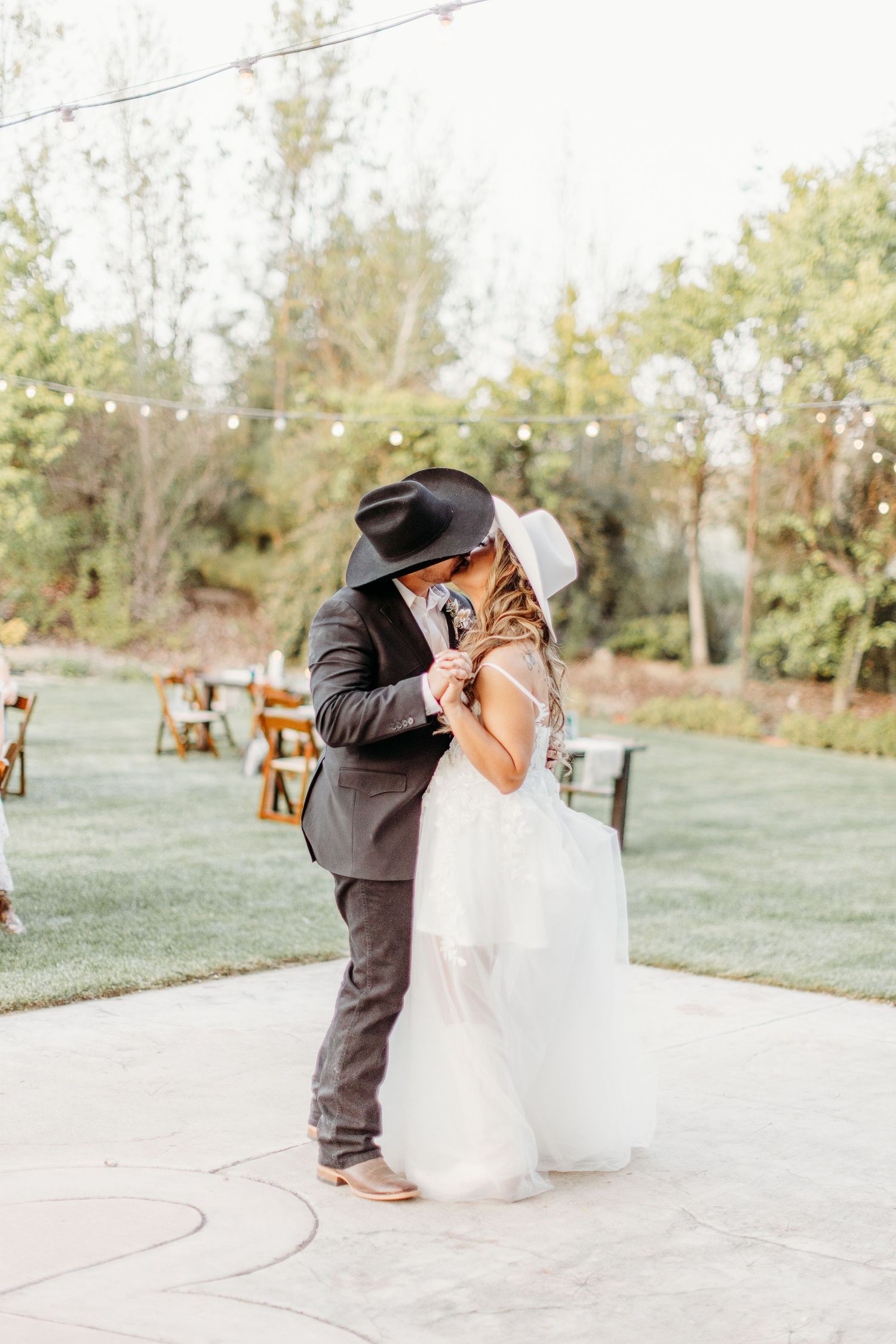 A couple shares an intimate moment during their outdoor wedding ceremony on a paved patio with wooden chairs.