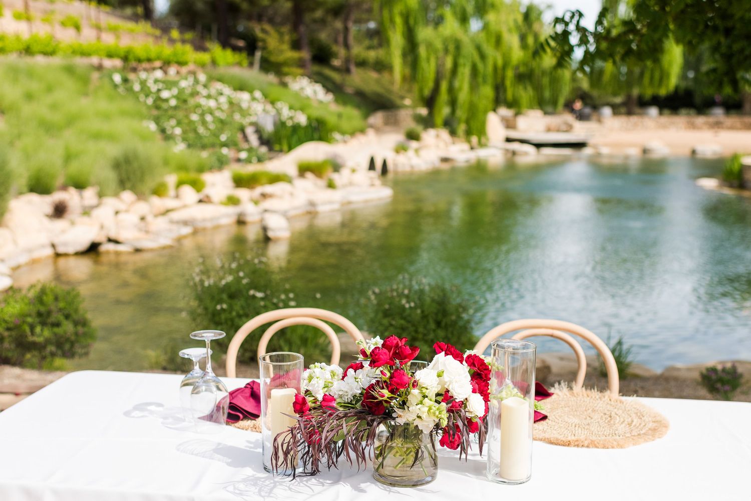 Pink and red floral centerpieces decorate a white table overlooking a serene pond with weeping willows.
