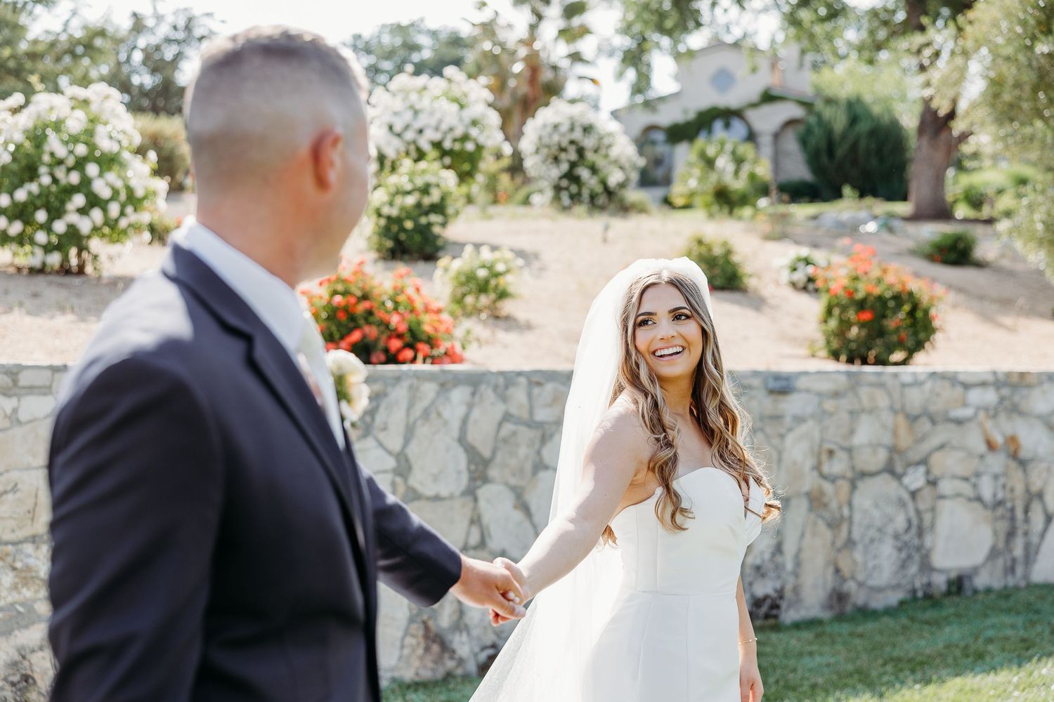 Newlyweds share a romantic moment on a garden path surrounded by stone walls and greenery during their wedding day photoshoot.