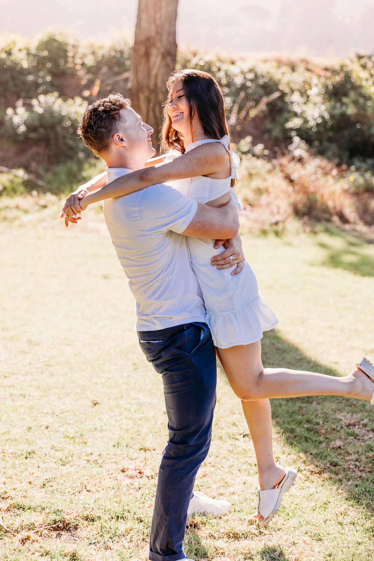 Playful couple in white outfits sharing joyful moment in sunlit garden.