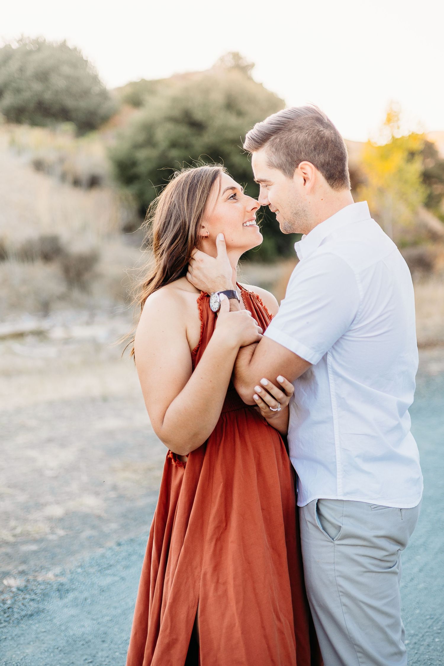 Tender moment between couple in white shirt and rust dress against natural landscape.