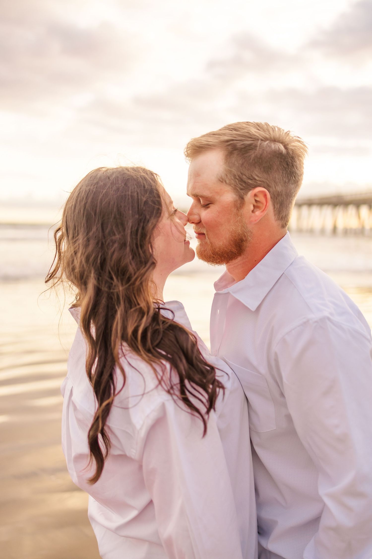 Intimate beachside moment with golden sunset light and ocean pier in background.