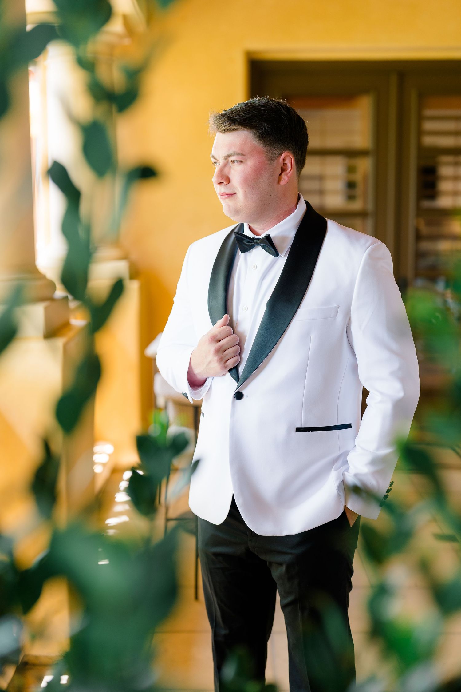 Groom in white tuxedo jacket with black bow tie standing near greenery.