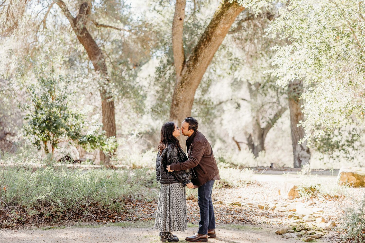 A couple shares a tender moment together in a sunlit outdoor setting with trees in the background.