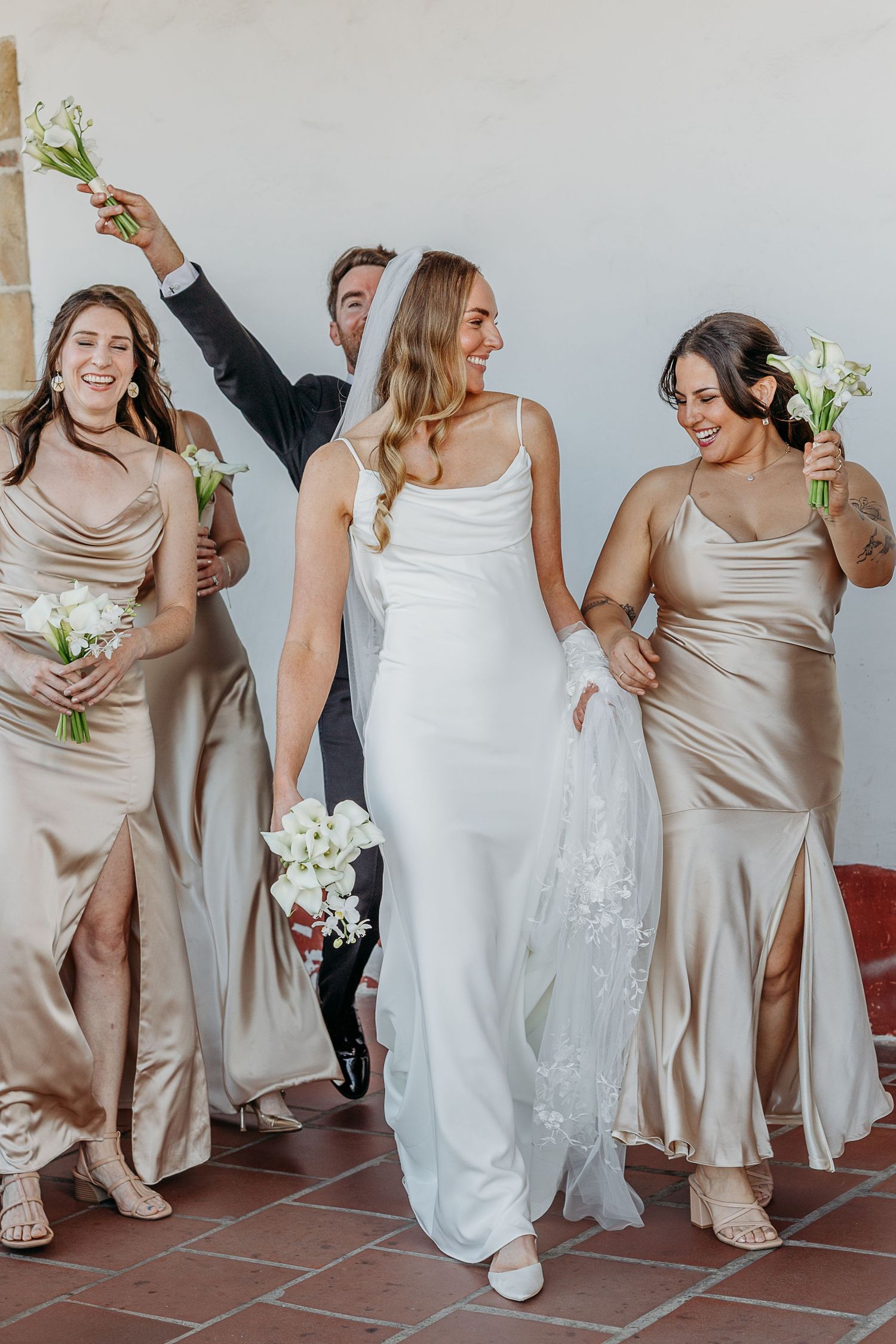Bridal party in champagne dresses and white gown laugh together during photo session.