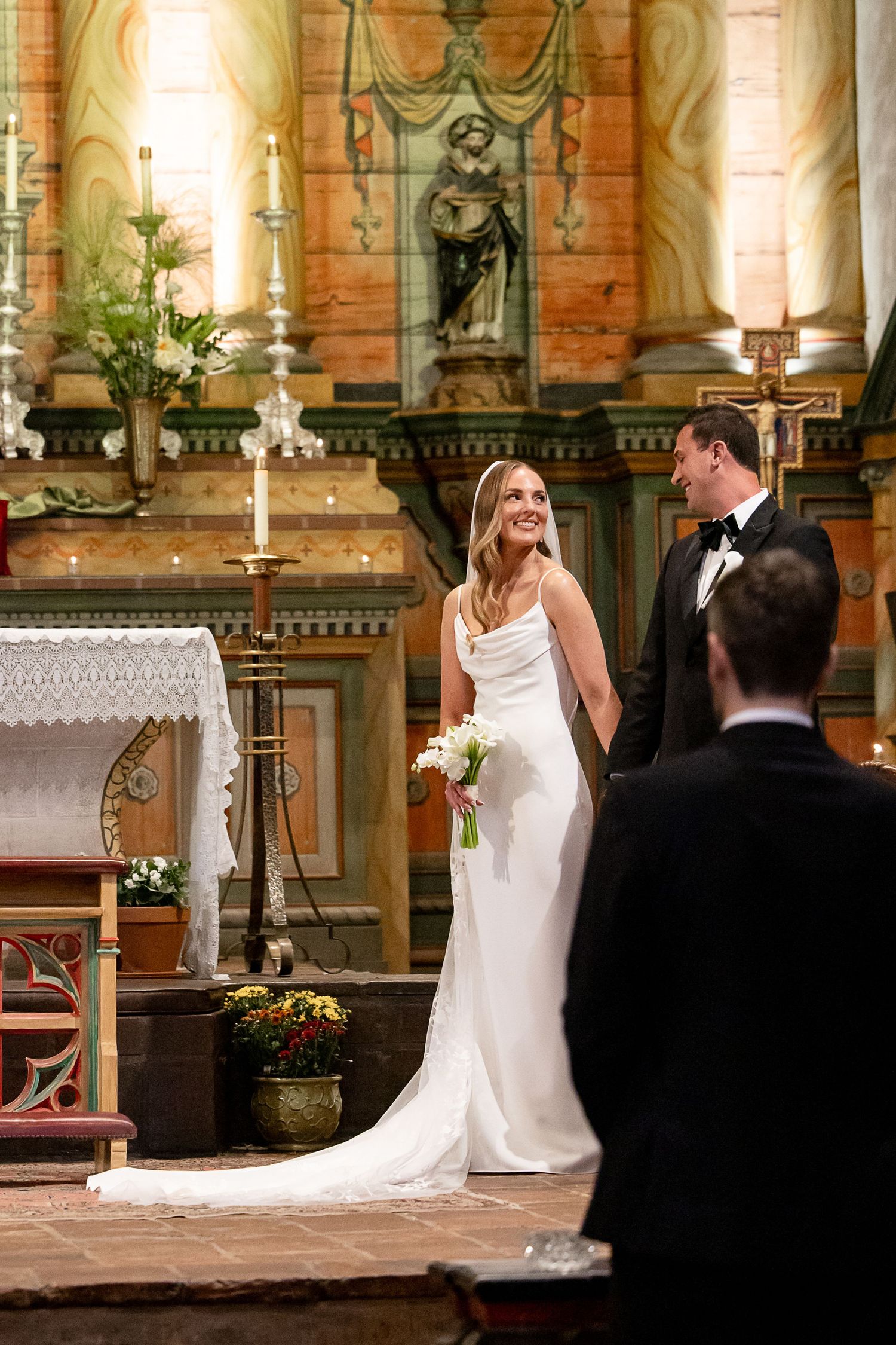 A bride in an elegant white fitted wedding dress stands at the altar of an ornate church during a ceremony.