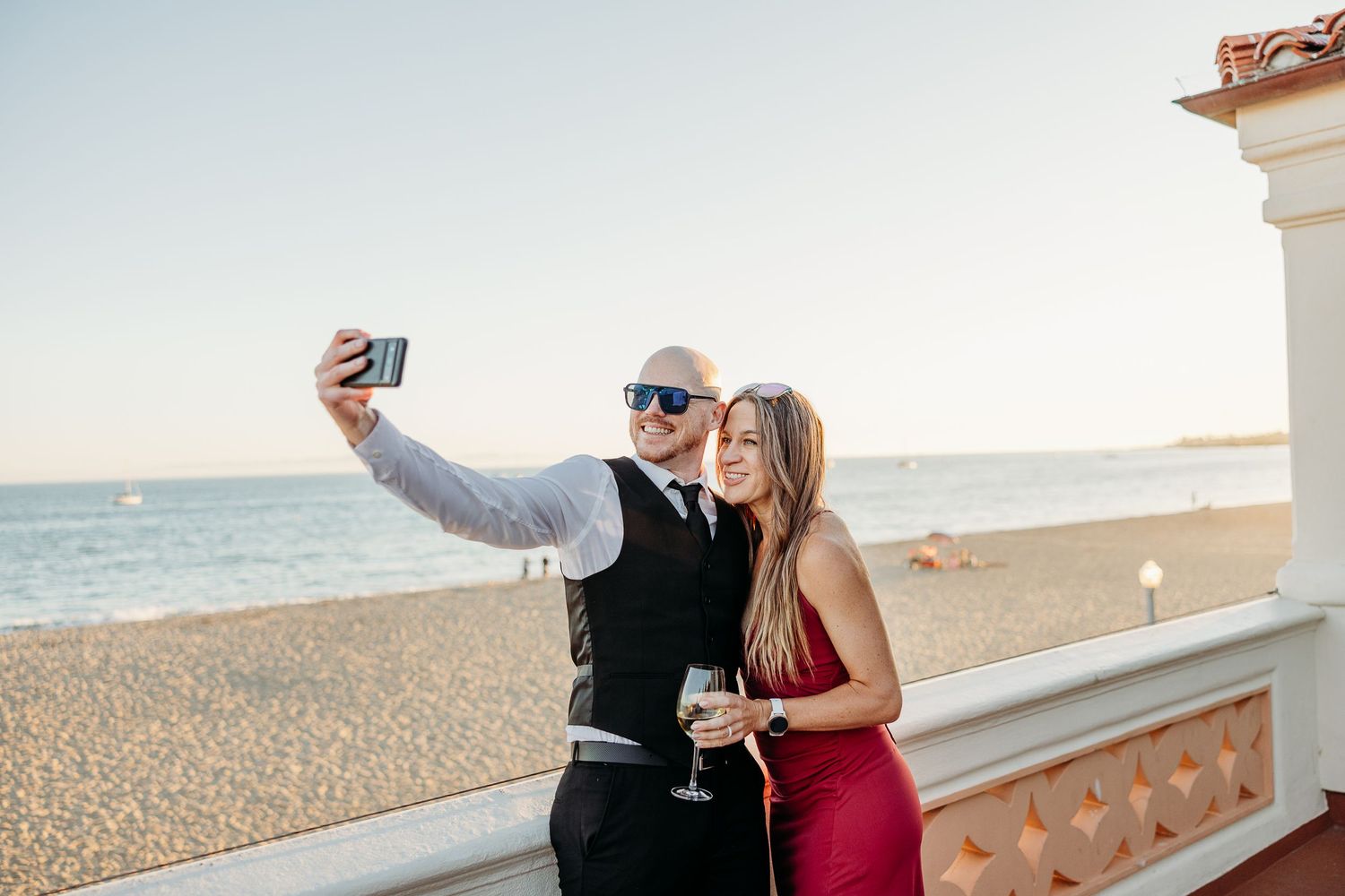 Couple taking selfies and enjoying drinks together on a beachfront terrace at sunset.