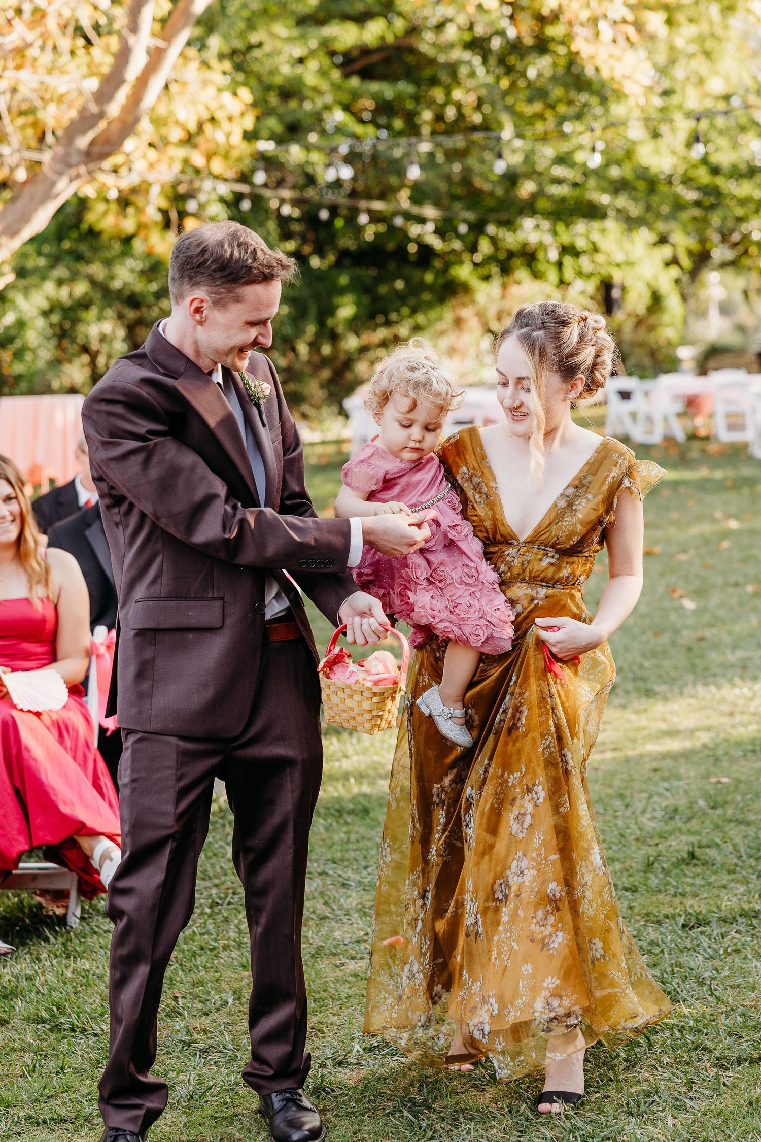 Outdoor wedding scene with people in formal attire interacting on grass with golden light.