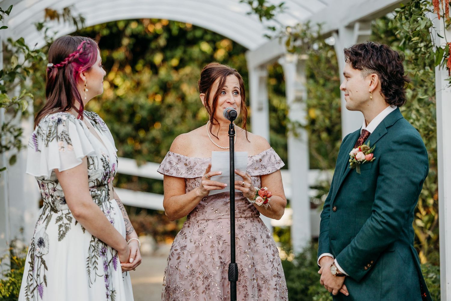 Outdoor wedding ceremony under a white pergola with guests seated on white chairs.