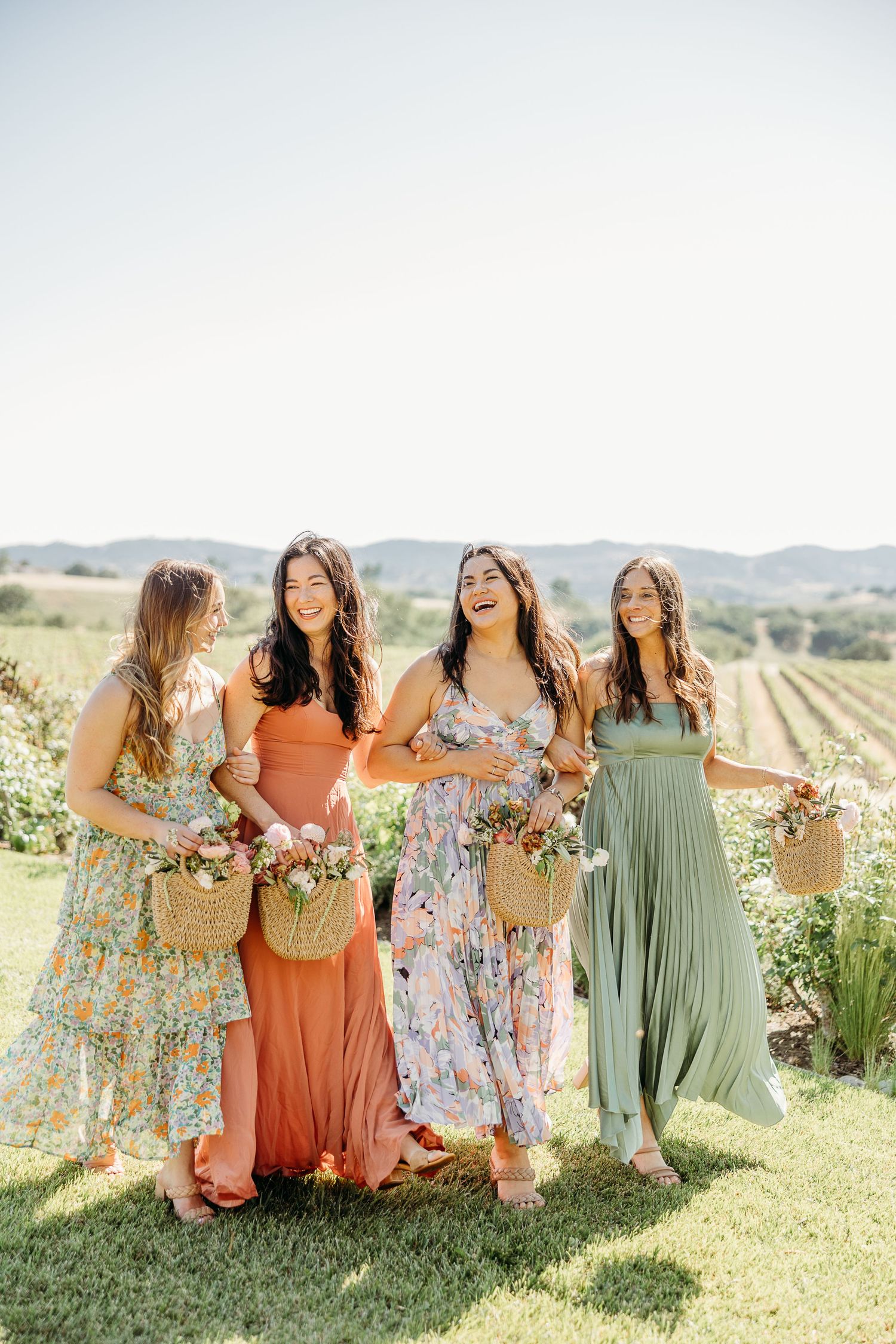 Bridesmaids in floral and solid dresses laugh together in a sunny vineyard field.
