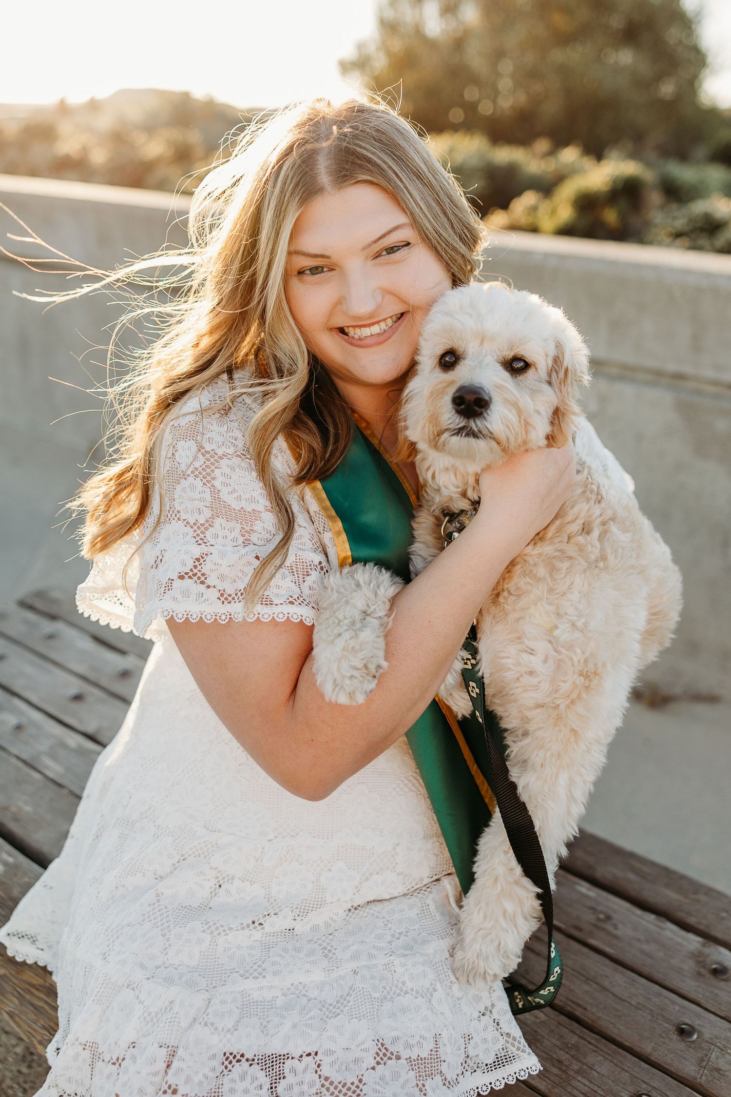 Bride in white lace dress hugs small white dog during outdoor wedding photo session.