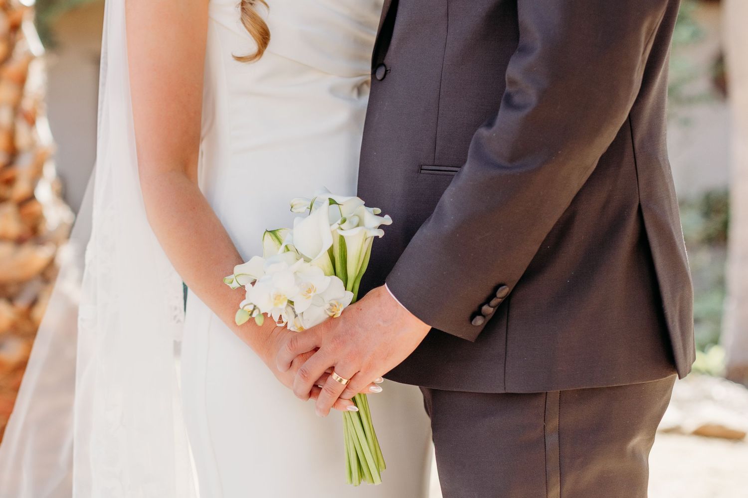 Bride and groom hold hands during an outdoor wedding ceremony while wearing elegant formal attire.
