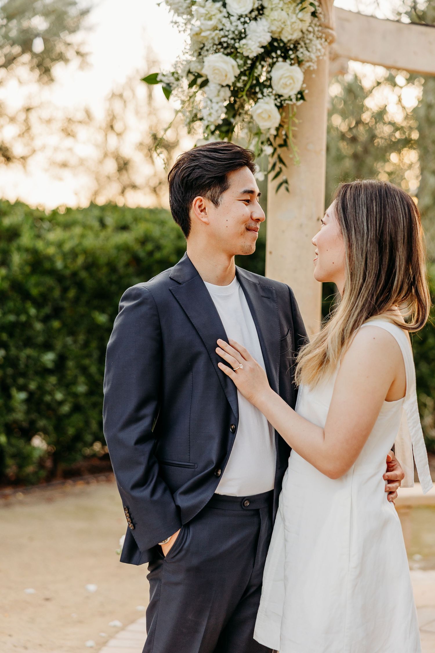 A couple shares an intimate moment under a garden arch decorated with white flowers at golden hour.