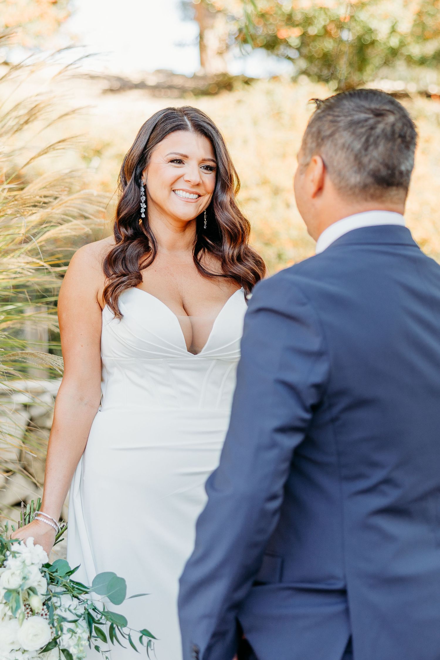 A couple in wedding attire poses together in a field of tall grass during golden hour.