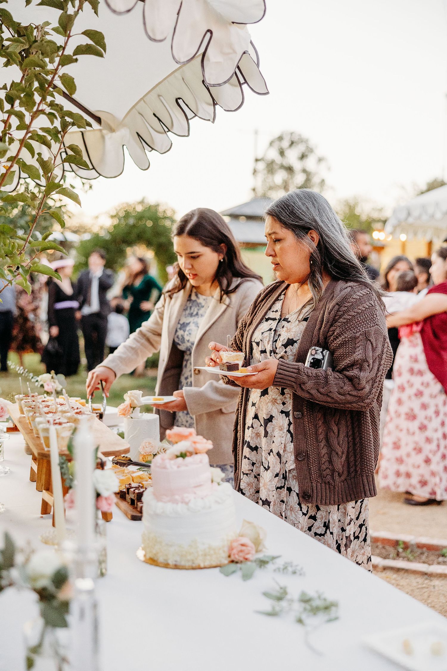 Wedding guests gather around dessert table with cake at outdoor reception during sunset.