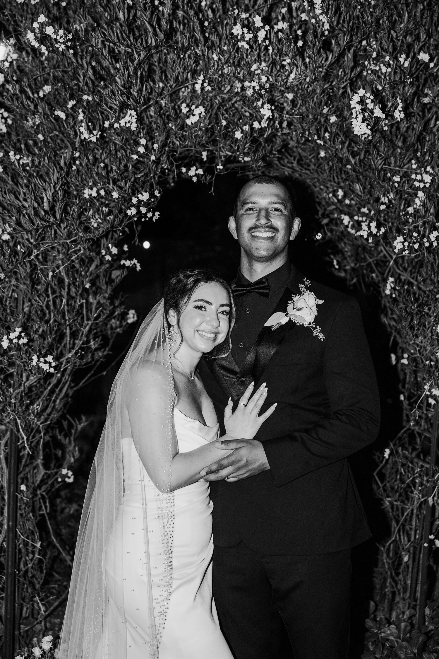 A black and white portrait of newlyweds smiling against a backdrop of flowering branches.