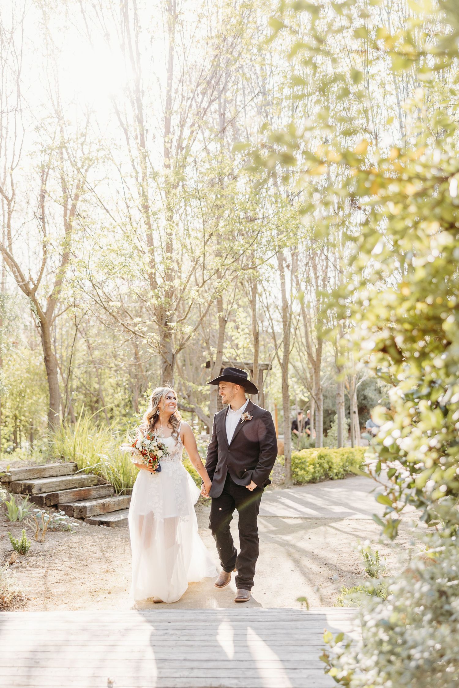 A couple walks along a sunlit woodland path during their wedding photoshoot.