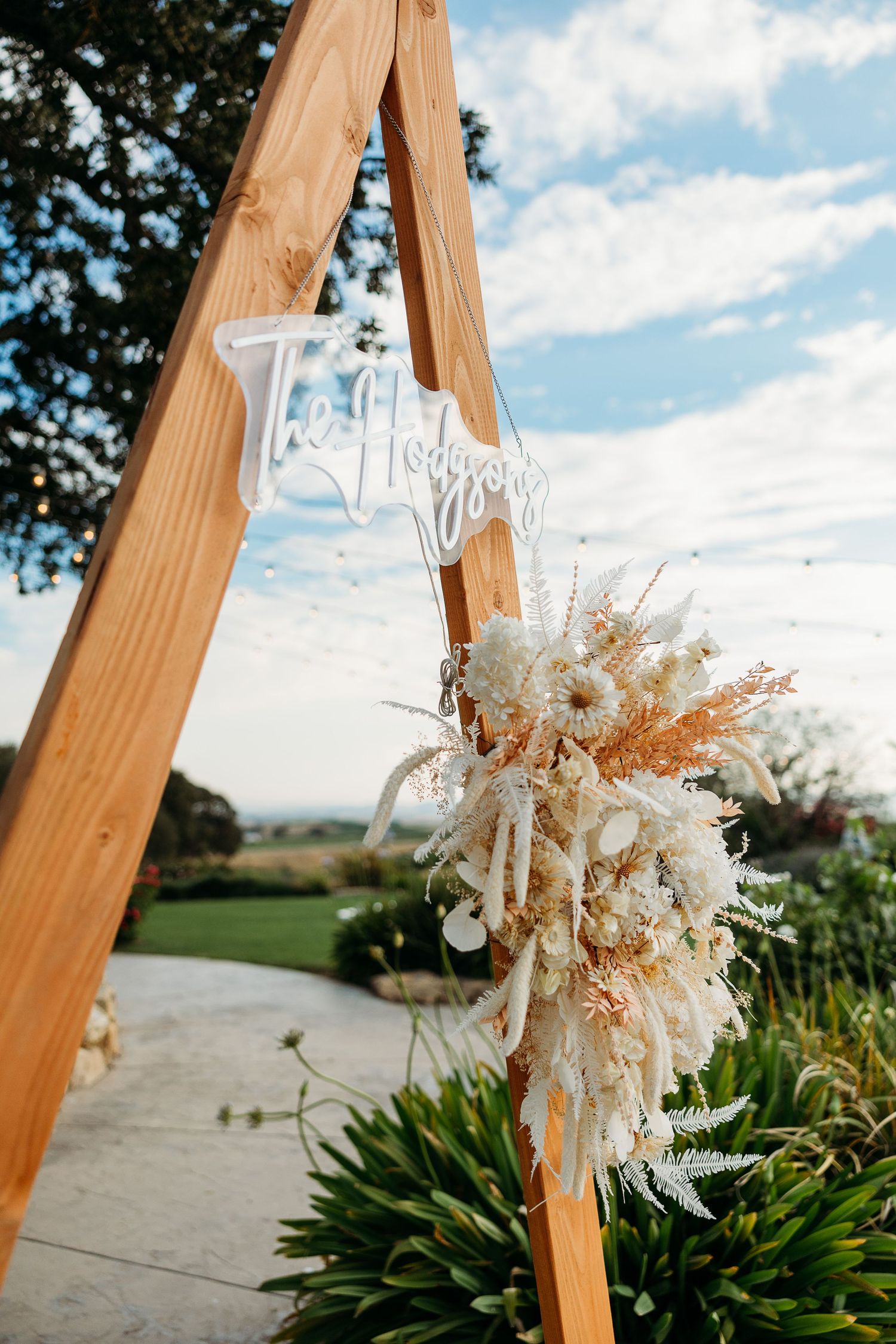 Wooden triangle arch with white neon sign and dried floral arrangement against blue sky.
