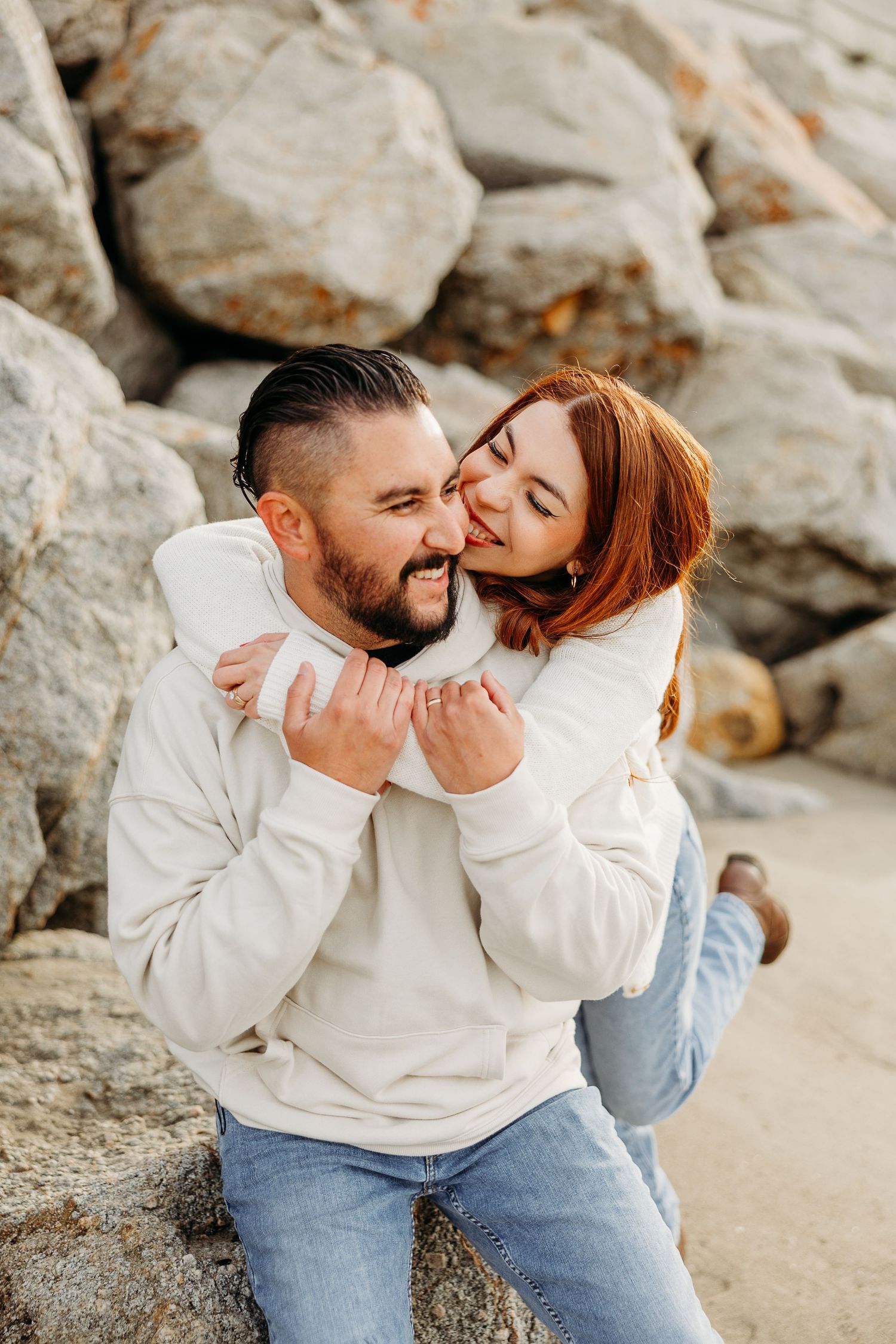 Two people in white sweaters and jeans sit close together on rocky beach terrain sharing intimate moments.