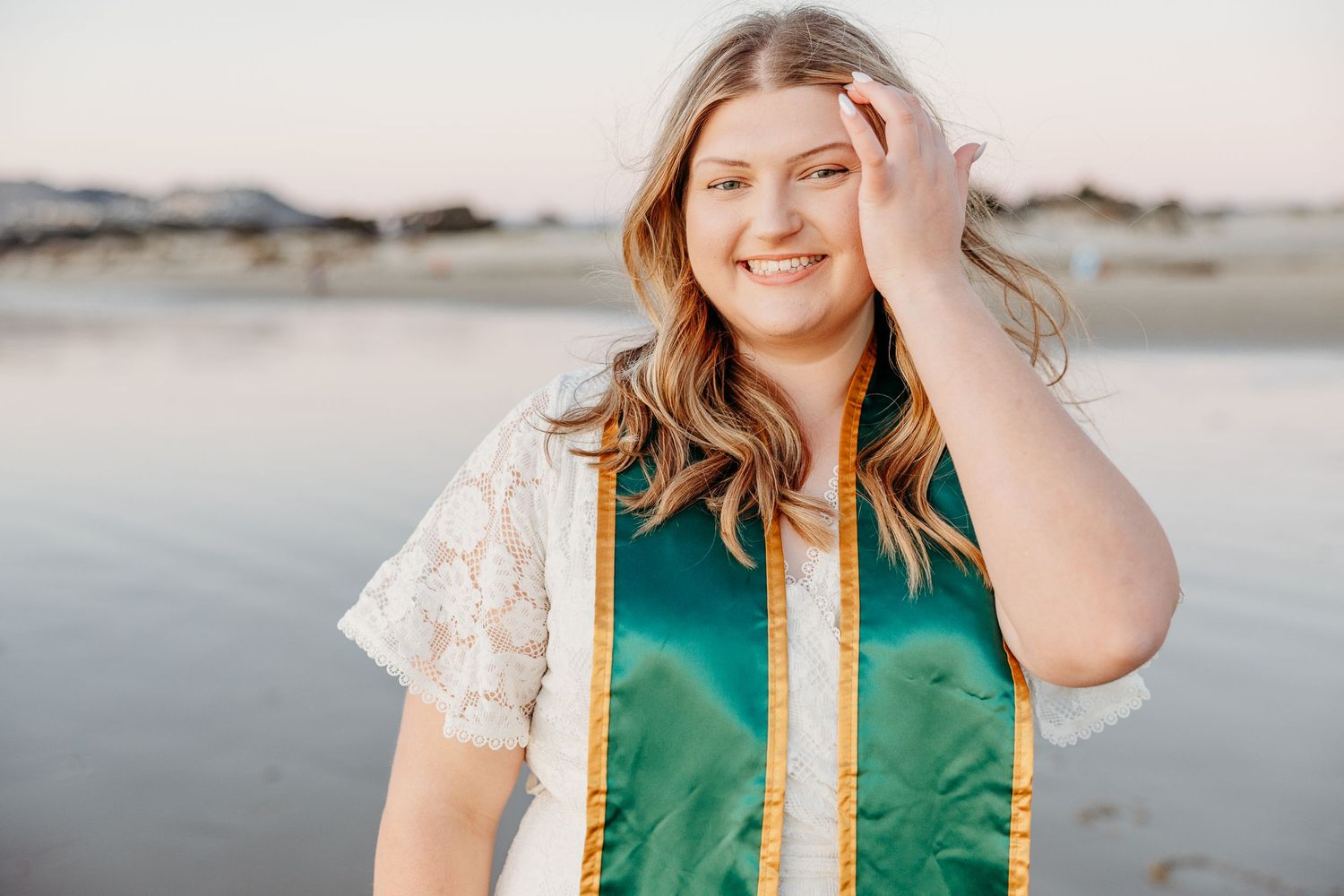Graduate in green sash and lace overlay smiles while posing at sunset near a beach.