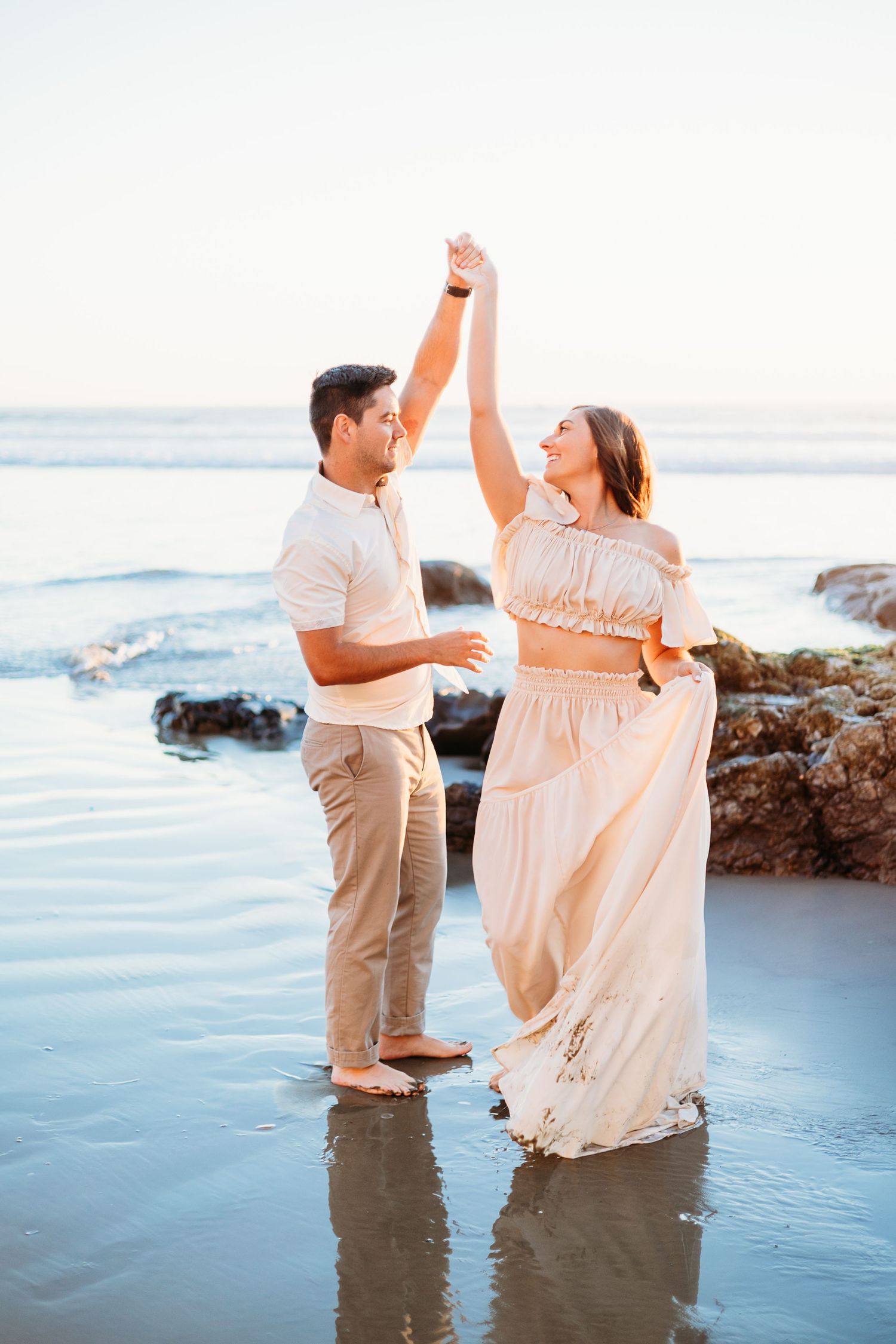 Dancing couple in flowing outfits splashing in ocean water at sunset near rocky shore.