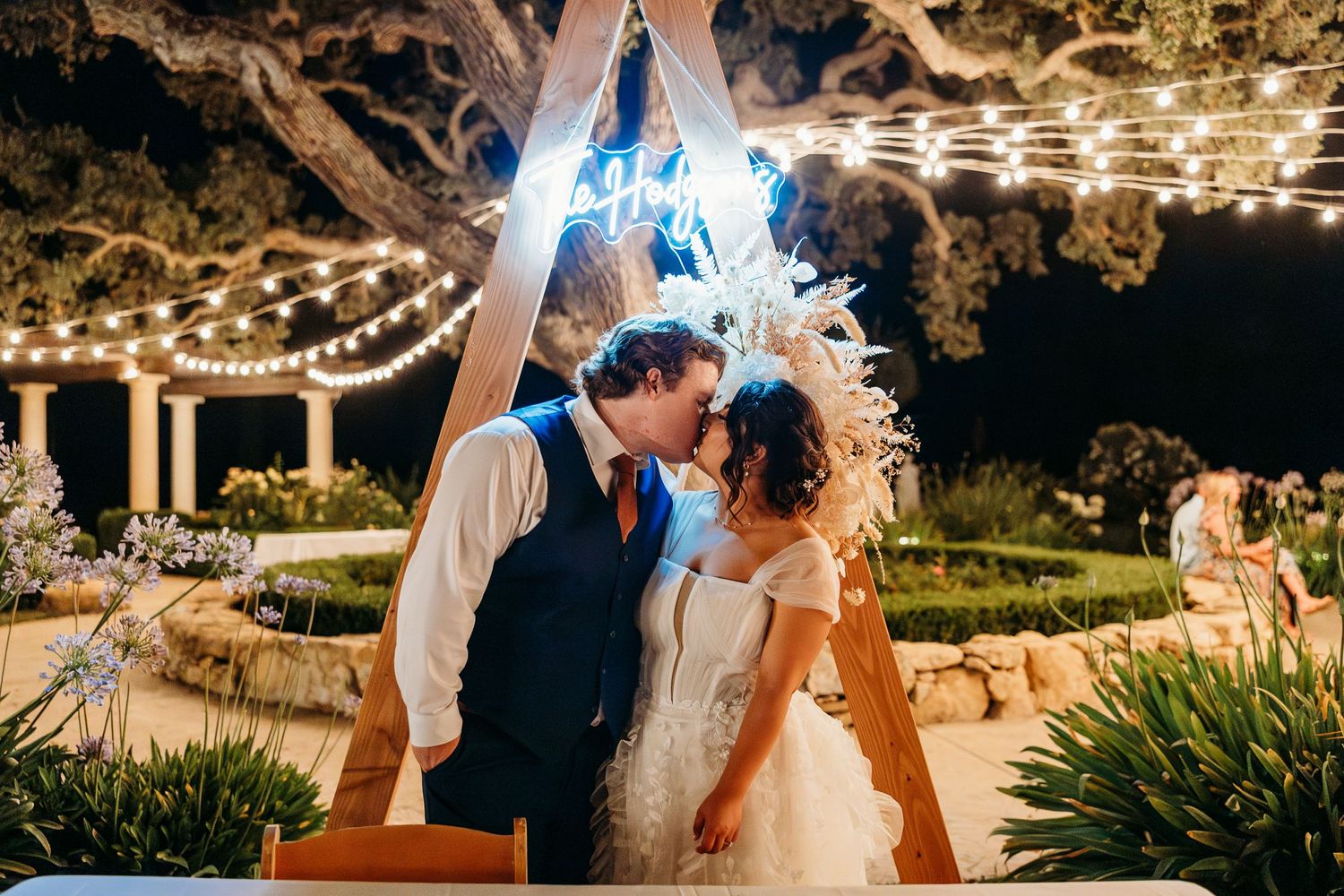 Couple sharing romantic moment under triangular arch with blue neon light and string lights.