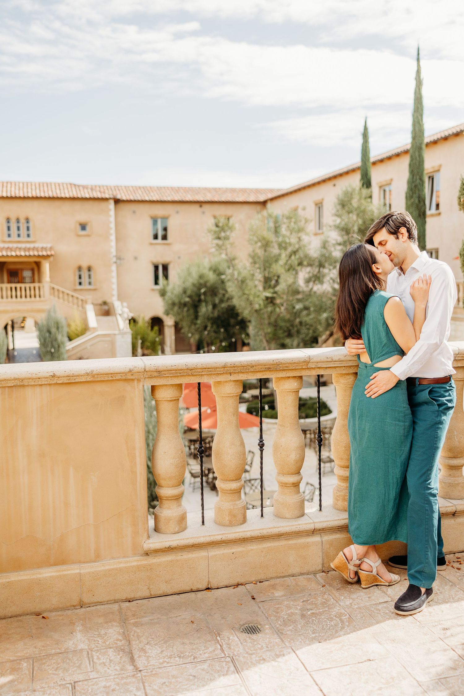 A romantic couple sharing a tender moment on a Mediterranean style balcony with beige architecture and cypress trees.