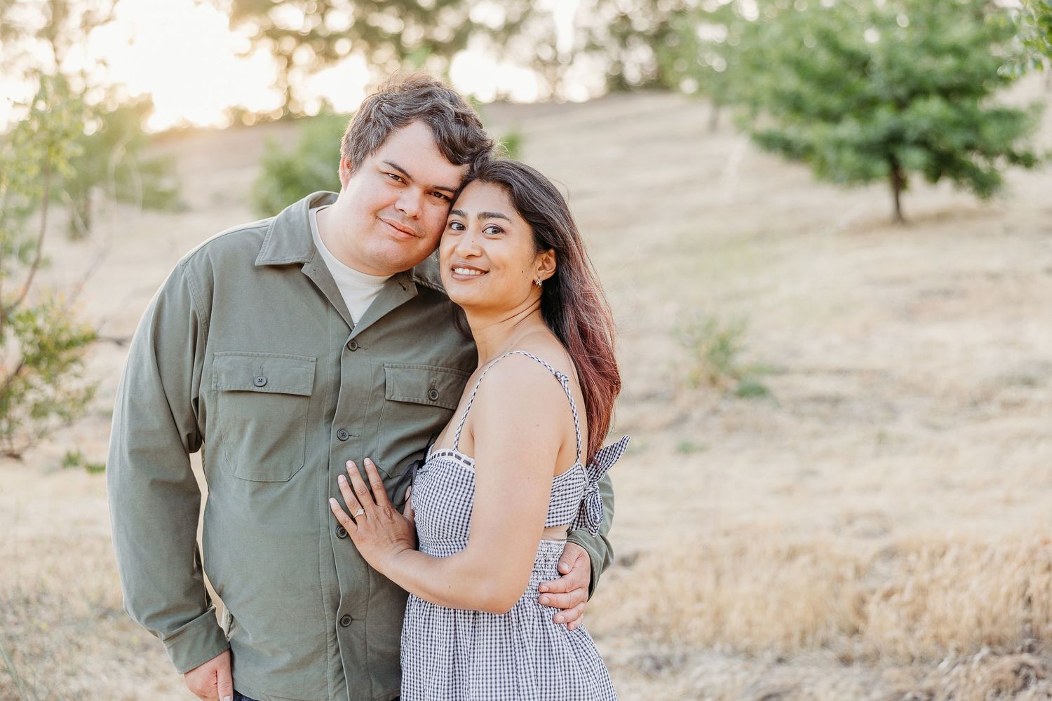 A couple embraces outdoors against a sunset background on a hillside with dried grass.