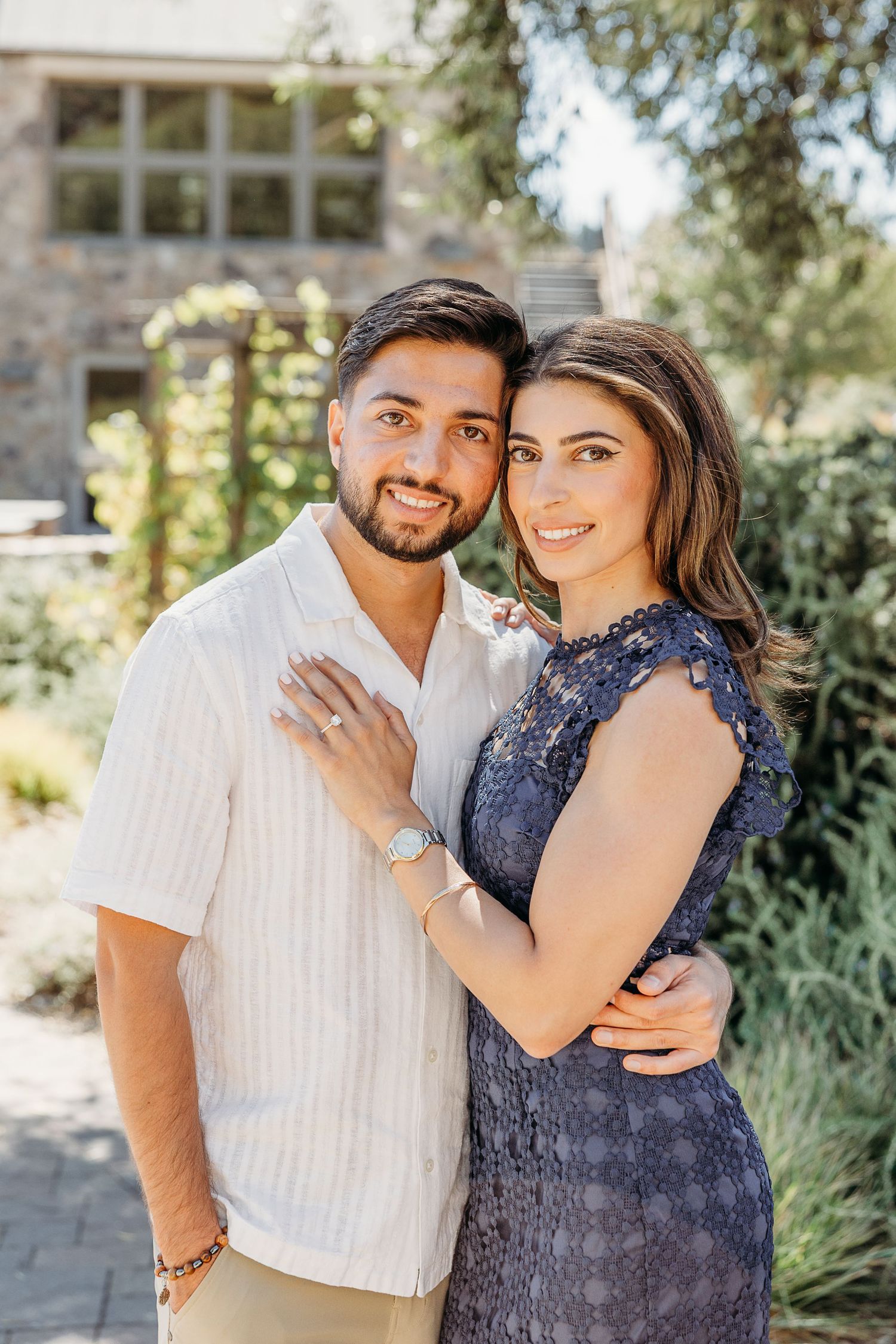 Couple embracing outdoors against a garden backdrop on a sunny afternoon.