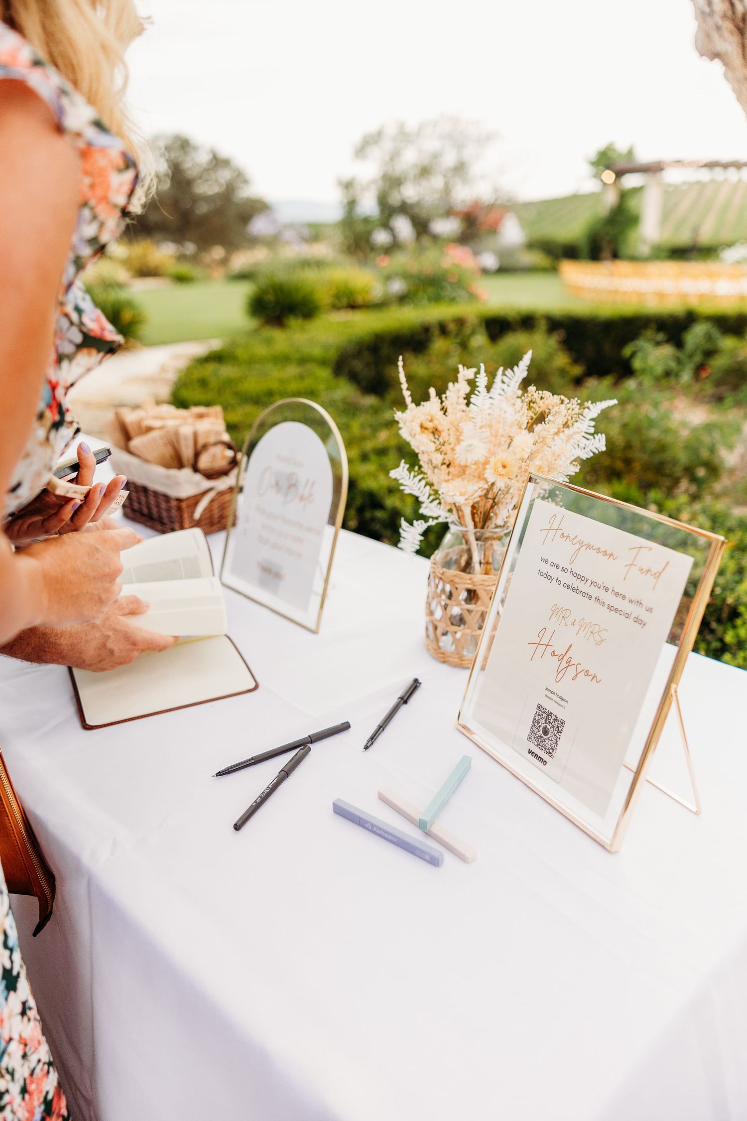 Wedding guest book table setup with dried flowers and framed signs at an outdoor venue.