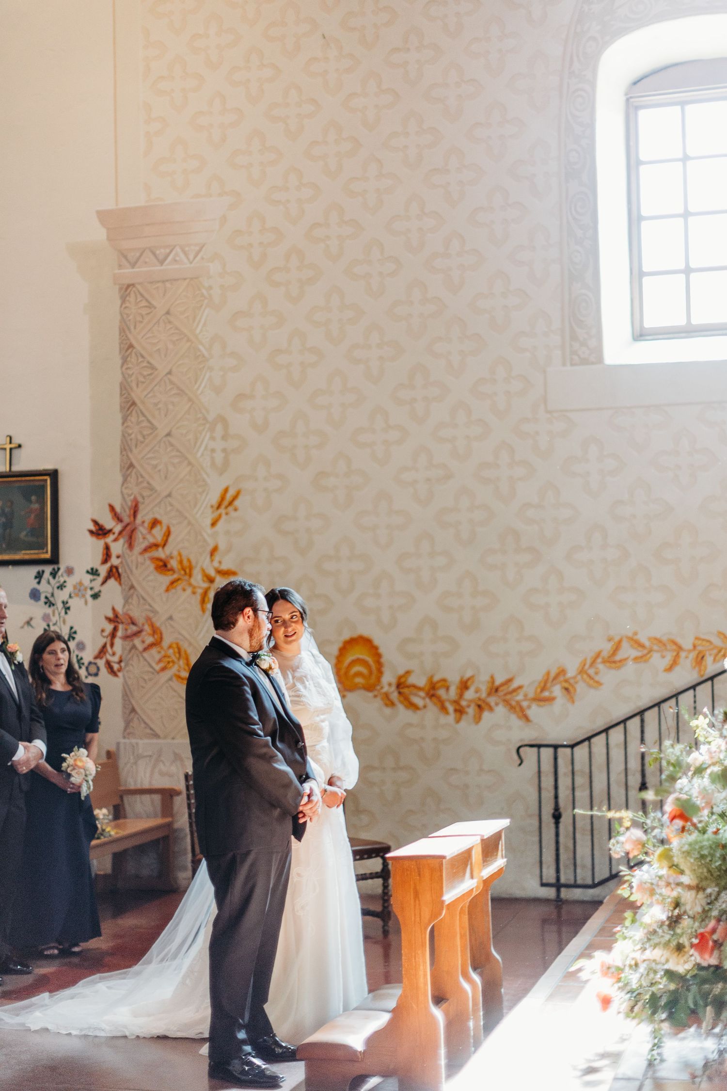 Wedding ceremony in an elegant white church with ornate wall patterns and soft natural lighting in Mission San Luis Obispo.