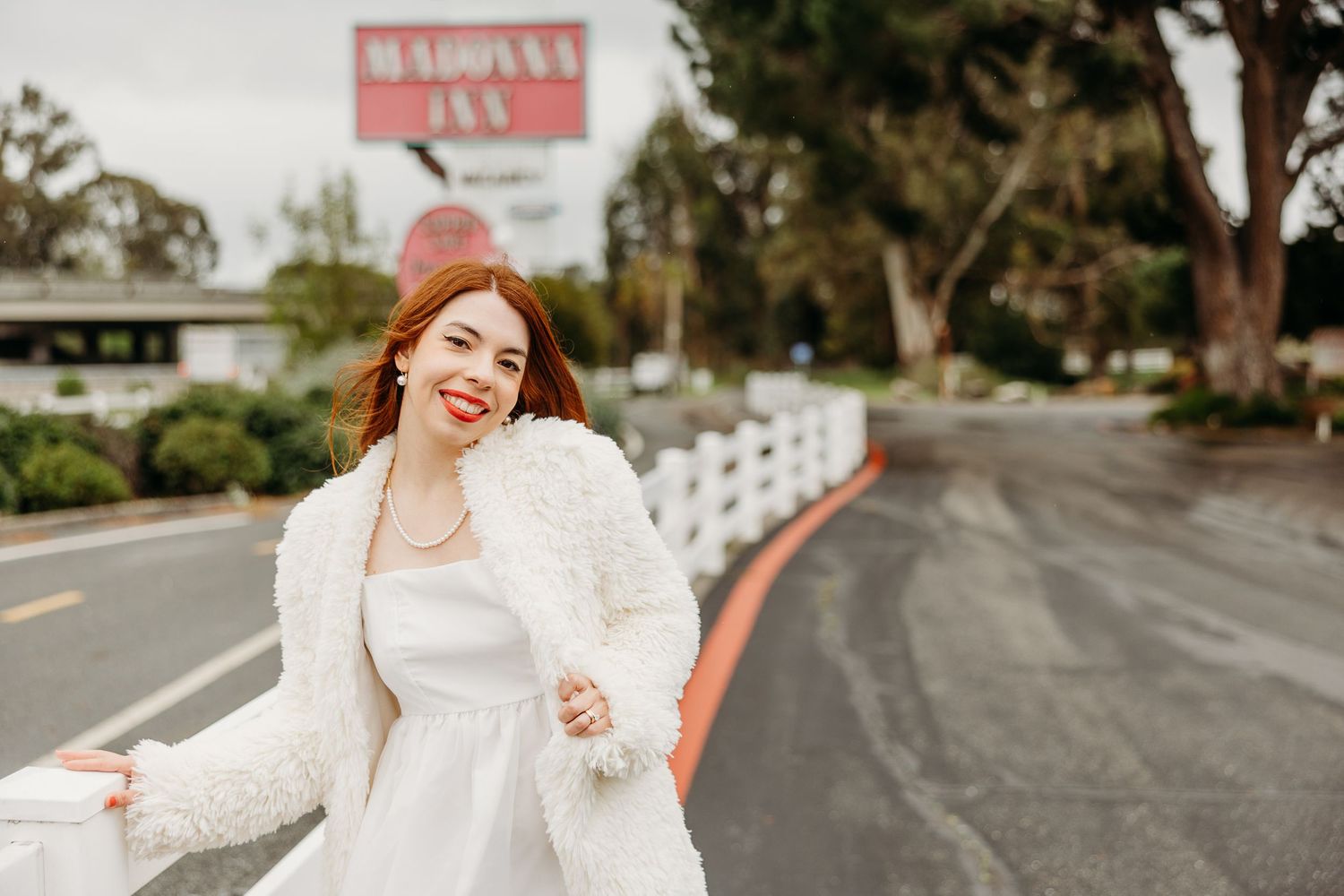 A person in a white furry coat walks along a curved road with trees and buildings in the background.