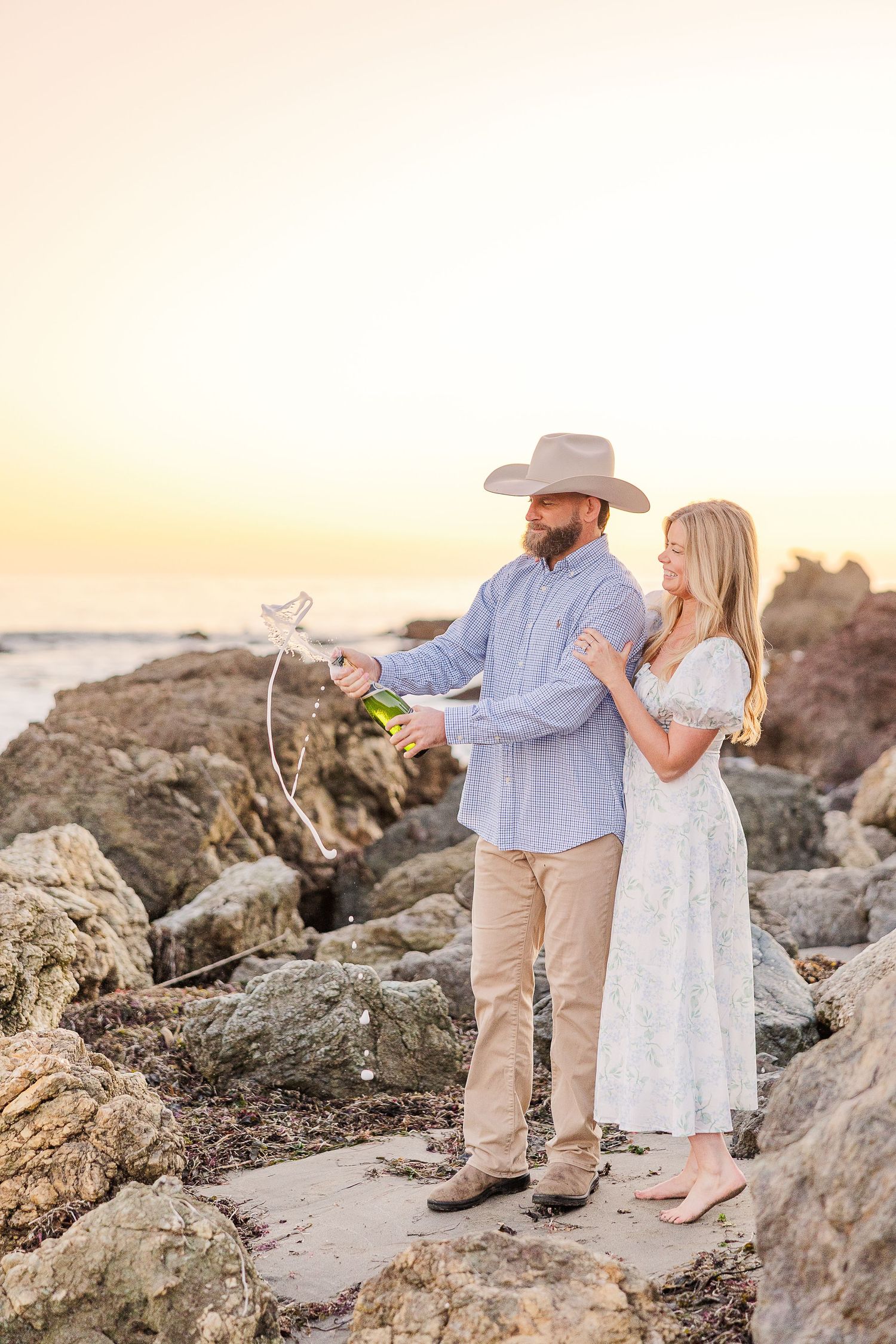 A couple holds champagne glasses while standing on coastal rocks at sunset.