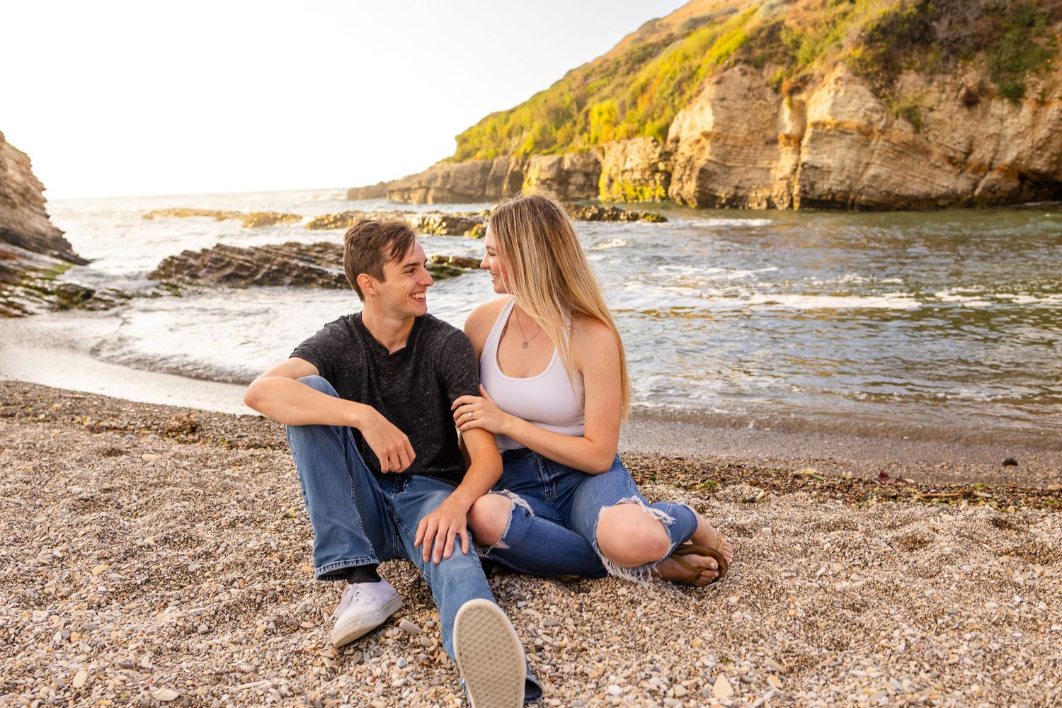 A couple sits casually together on a rocky beach during a sunny afternoon.
