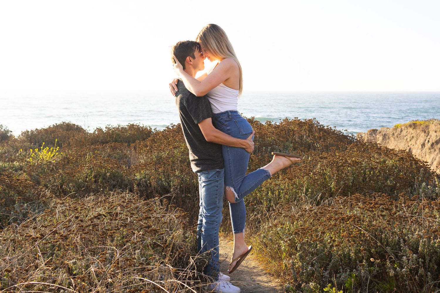 A couple shares an intimate embrace on a coastal trail during golden hour.