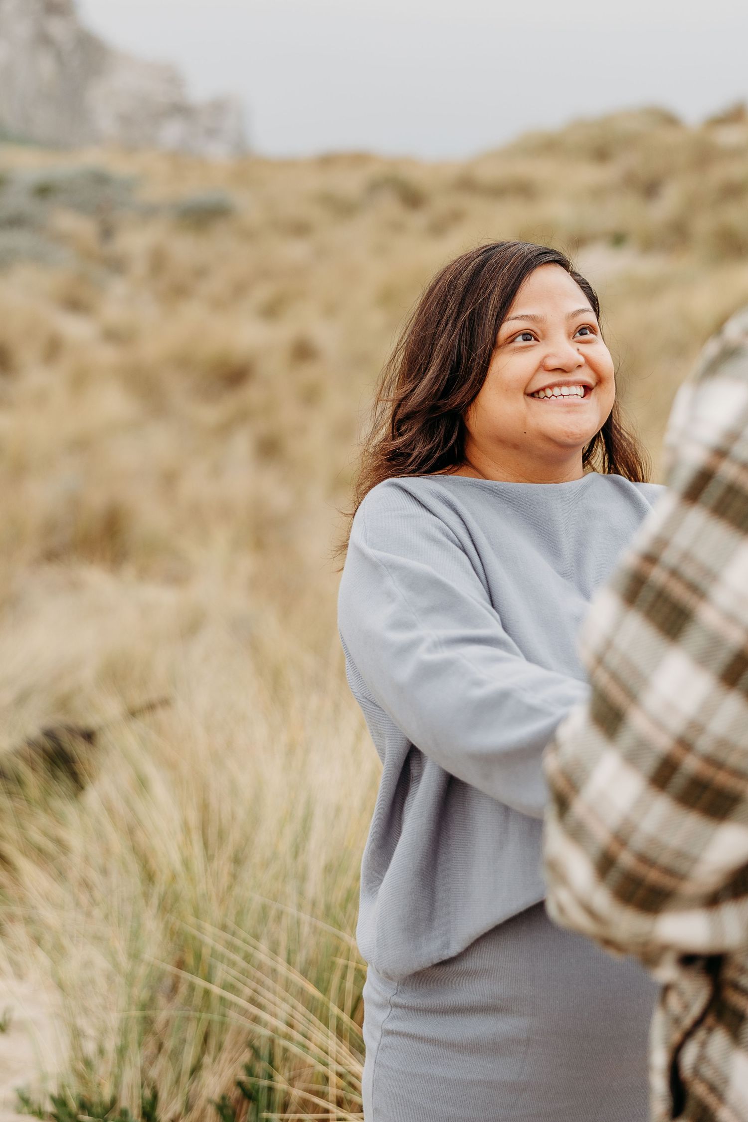 A smiling person wearing a gray sweater poses in a field of tall grass on a sunny day.