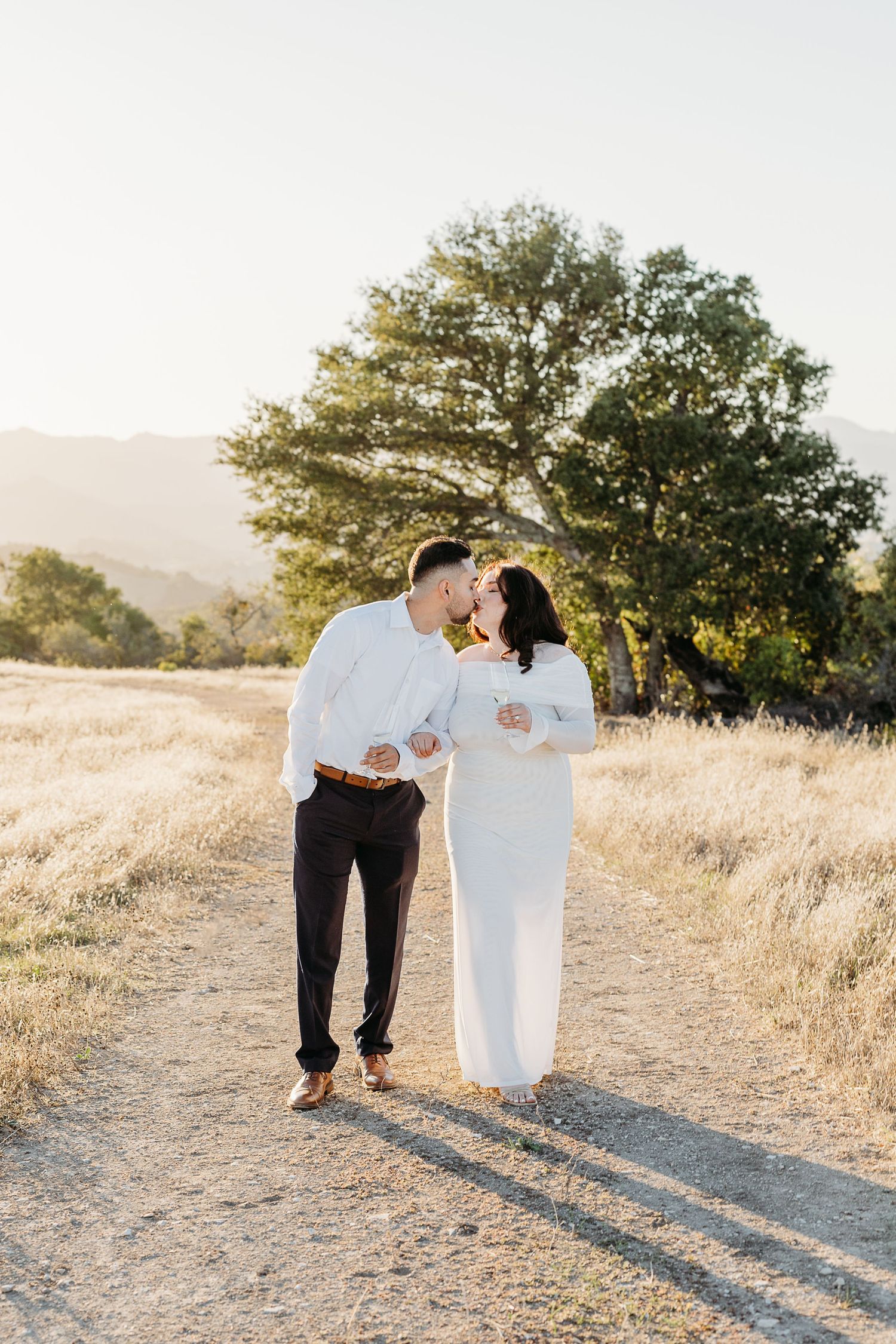 A couple shares an intimate moment on a sunlit dirt path surrounded by golden grass and trees at sunset.