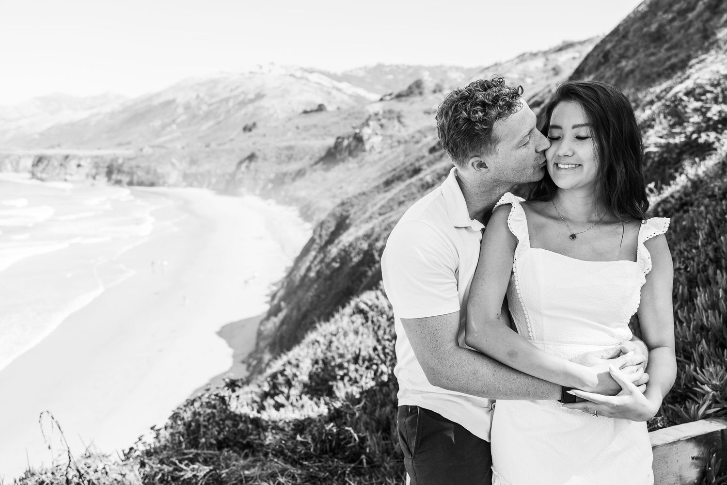 A black and white portrait of a couple sharing an intimate moment by the ocean.