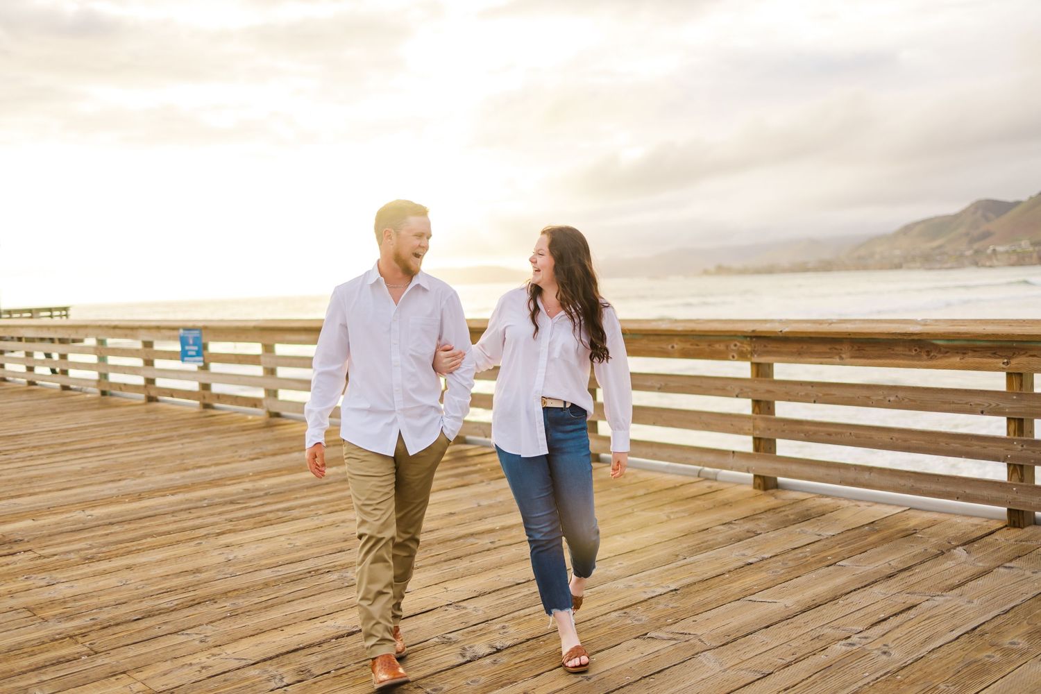 A couple walking hand in hand on a wooden pier with coastal views in the background.