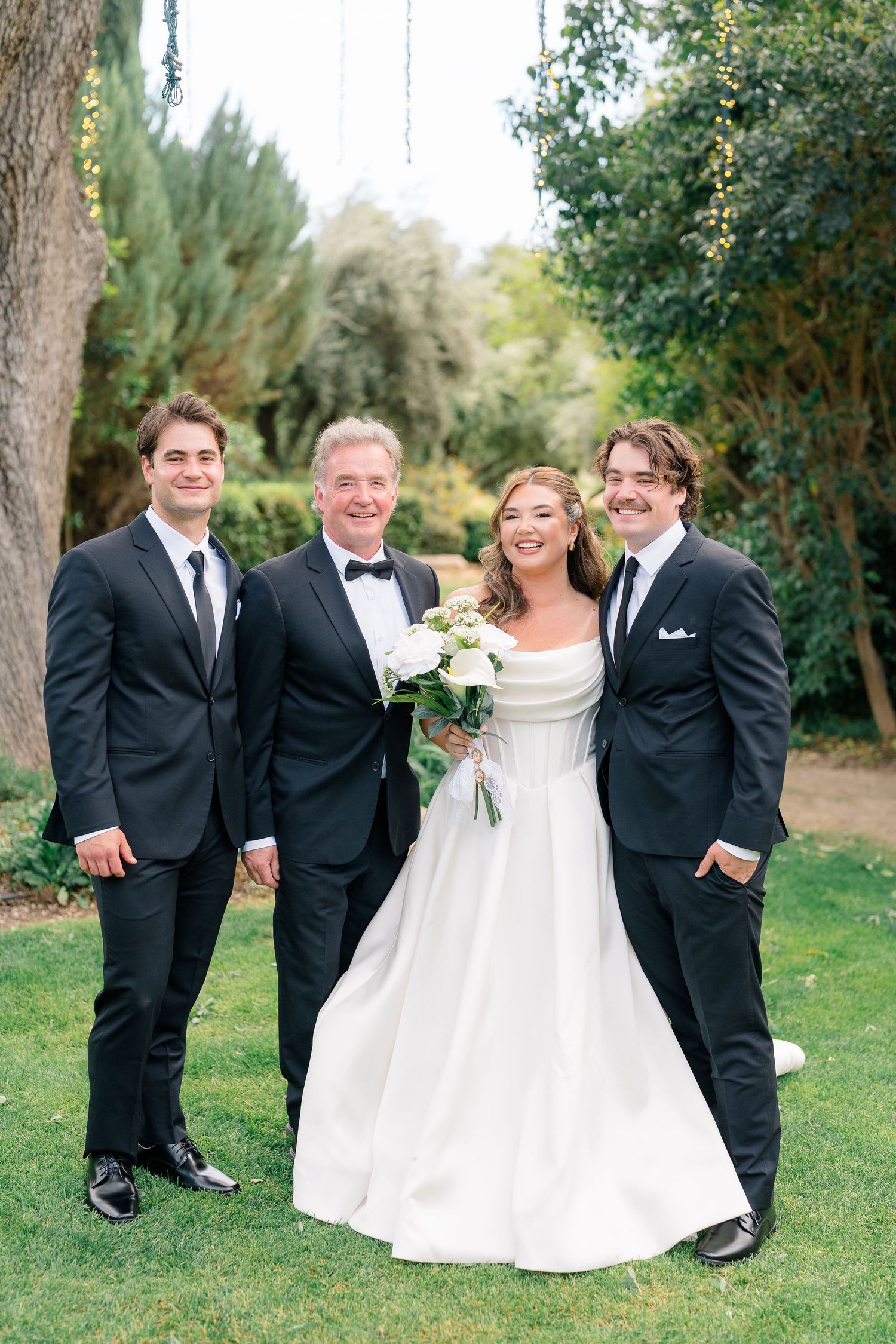 Bride in elegant white gown poses with three men in black tuxedos on green lawn at outdoor wedding.