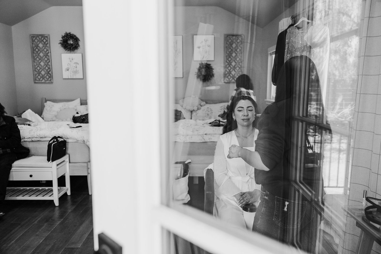 Black and white photo of a bride in a white robe preparing in a cozy bedroom, reflected in a window.