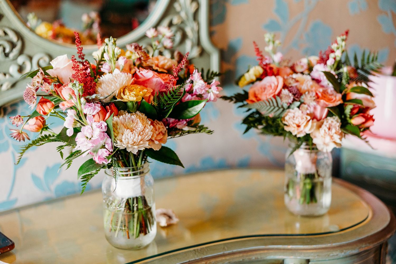 Two glass vases filled with vibrant pink and coral floral arrangements sit on a round table with blue wallpaper backdrop.